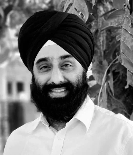 A man with a turban and beard smiling outdoors with leafy background.