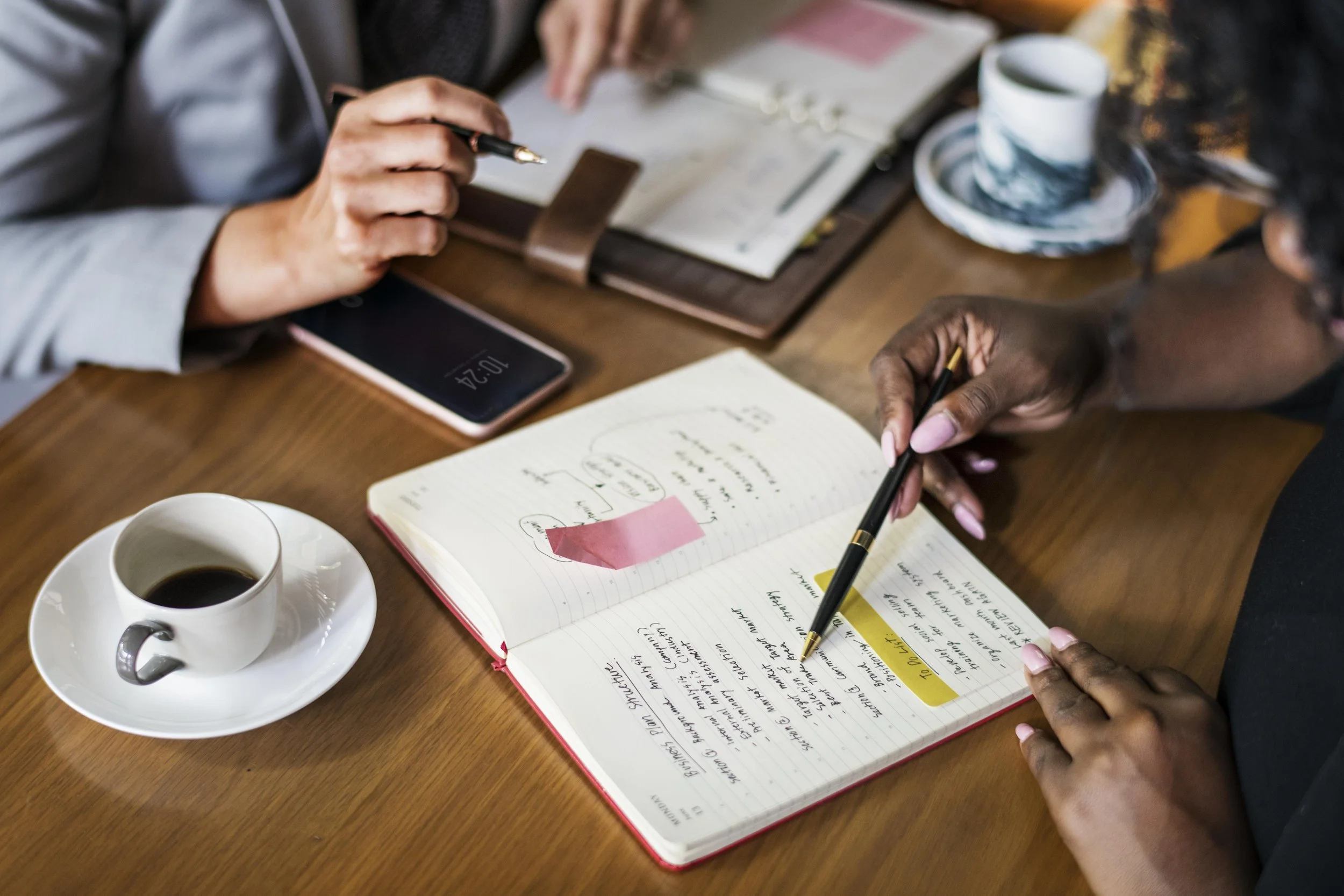 Two people sitting at a wooden table, engaged in a discussion, with a coffee cup, a smartphone, a notebook filled with handwritten notes, and a cup and saucer on the table.