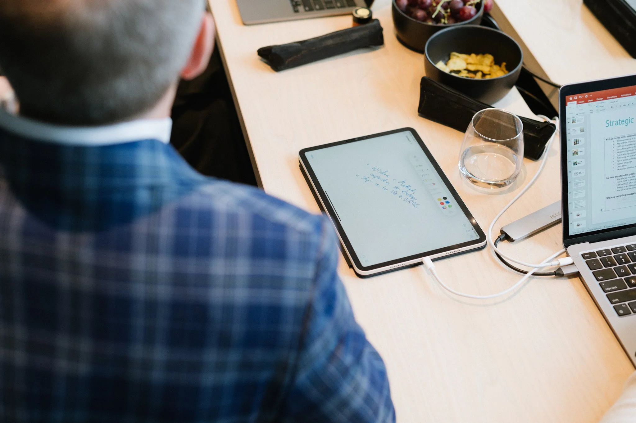 Overhead view of a person in a blue checkered shirt seated at a desk with a tablet, laptop, and various bowls of snacks, including chips and grapes, in a work setting.