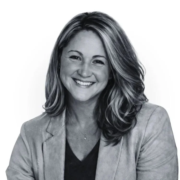 Black and white portrait of a smiling woman with shoulder-length wavy hair, wearing a blazer and a necklace.