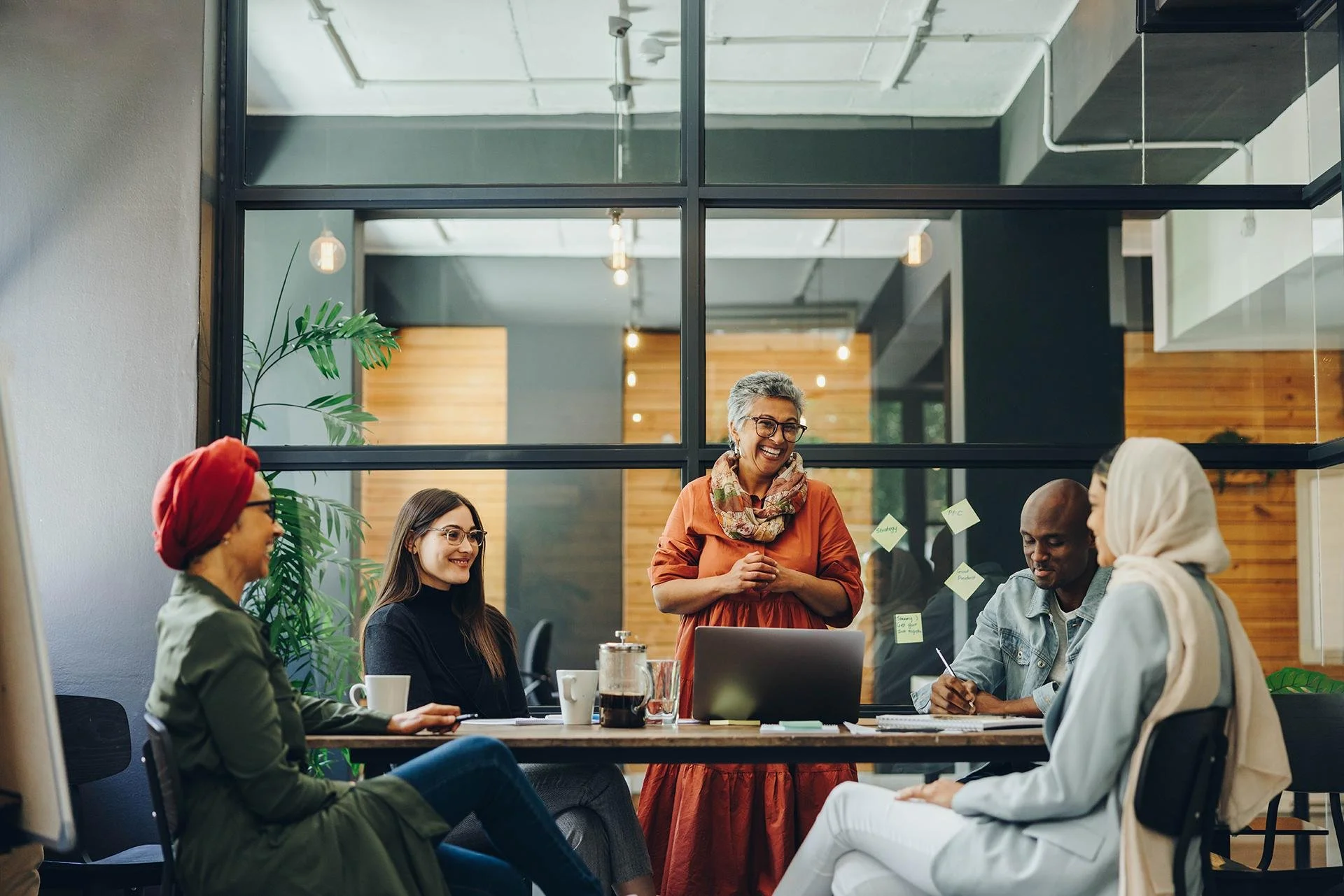 A diverse group of six people having a meeting in a modern office conference room, with a woman standing and leading the discussion. The group includes an older woman in an orange dress, two women and two men seated around the table. There are coffee cups, a laptop, and sticky notes on the glass wall behind them.