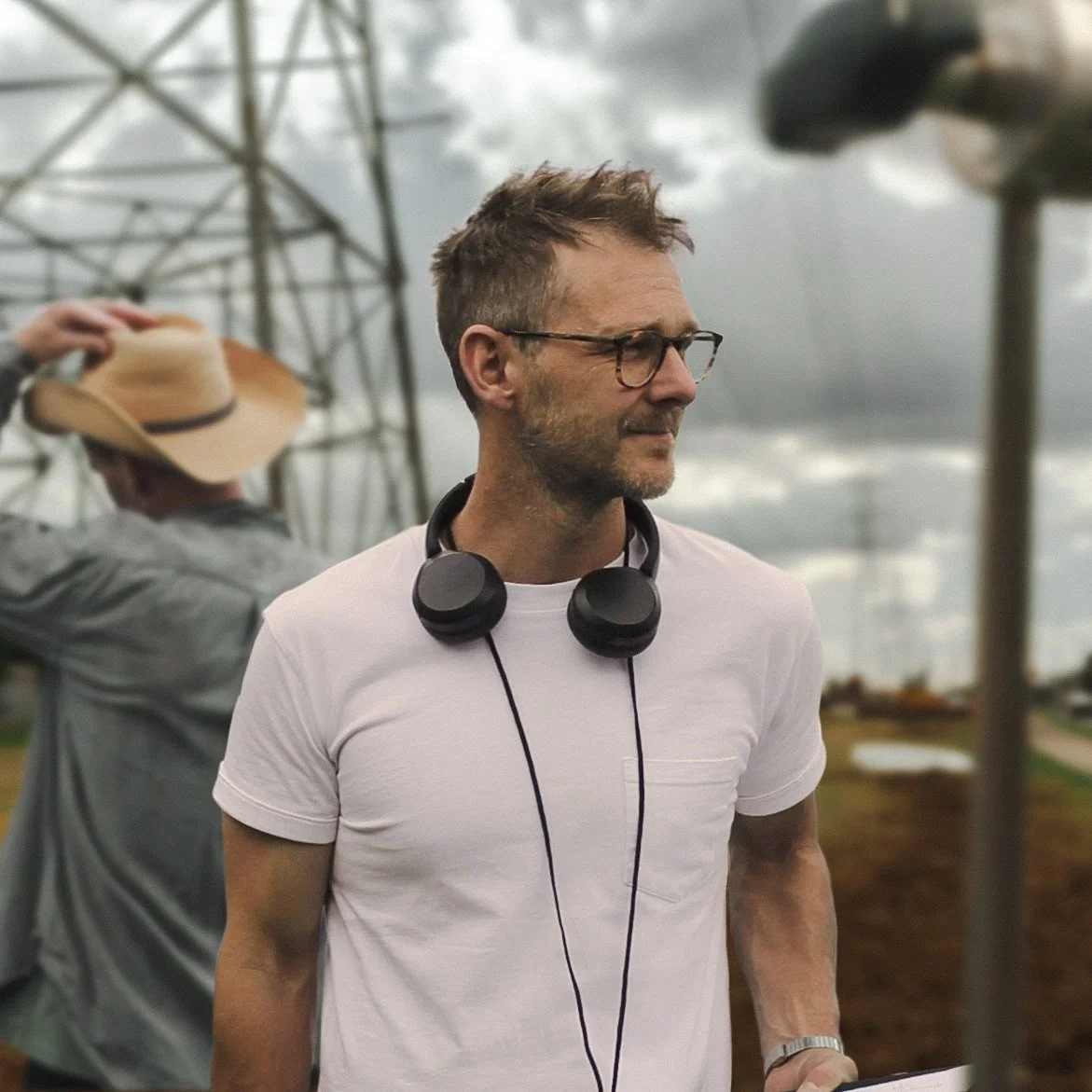 A man with glasses and headphones around his neck stands outdoors under a cloudy sky, wearing a white t-shirt.