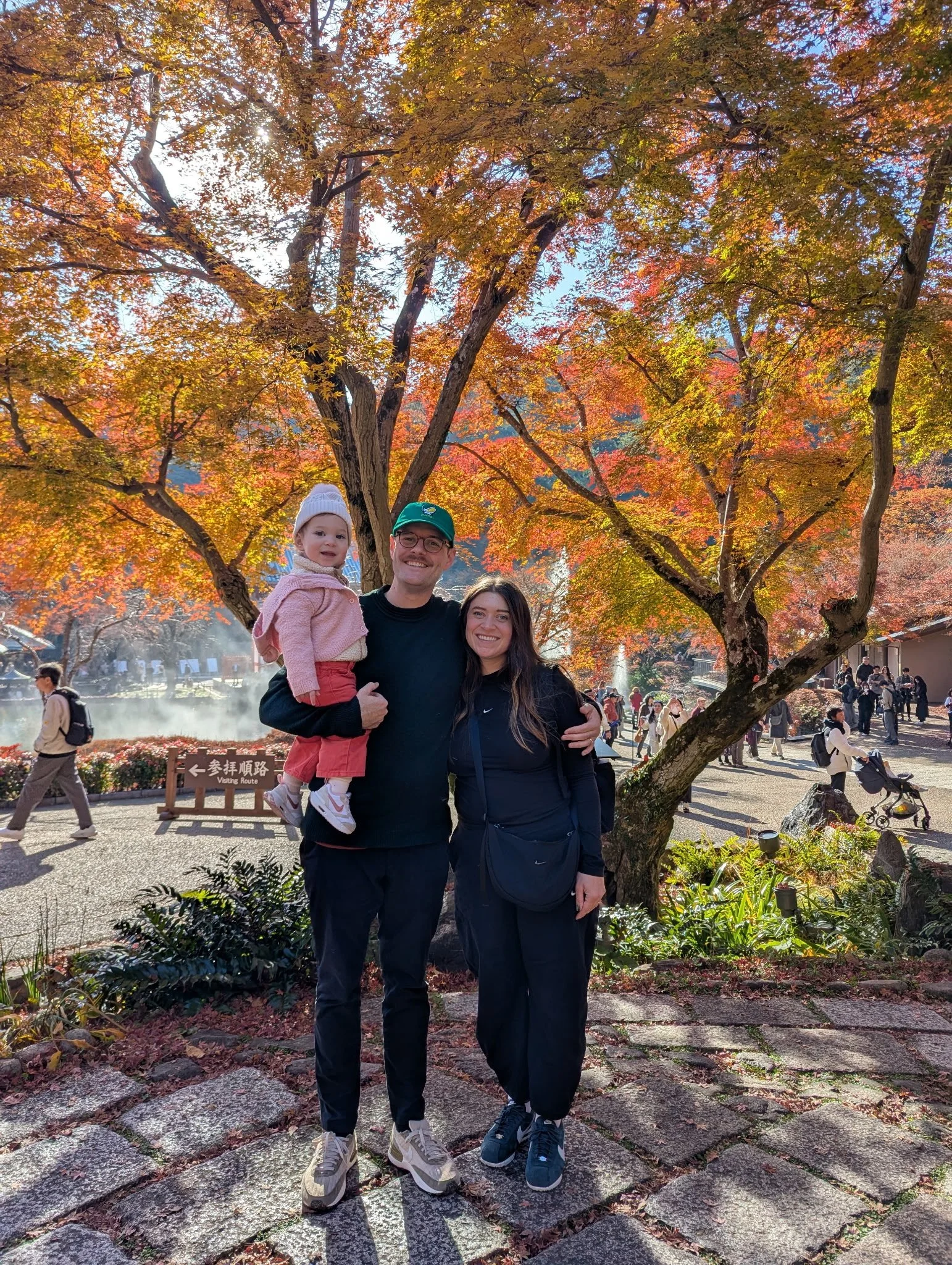 A family of three standing on a stone pathway under colorful autumn trees. The father is holding a young girl, and the mother is standing beside them. There are other people in the background enjoying the park.