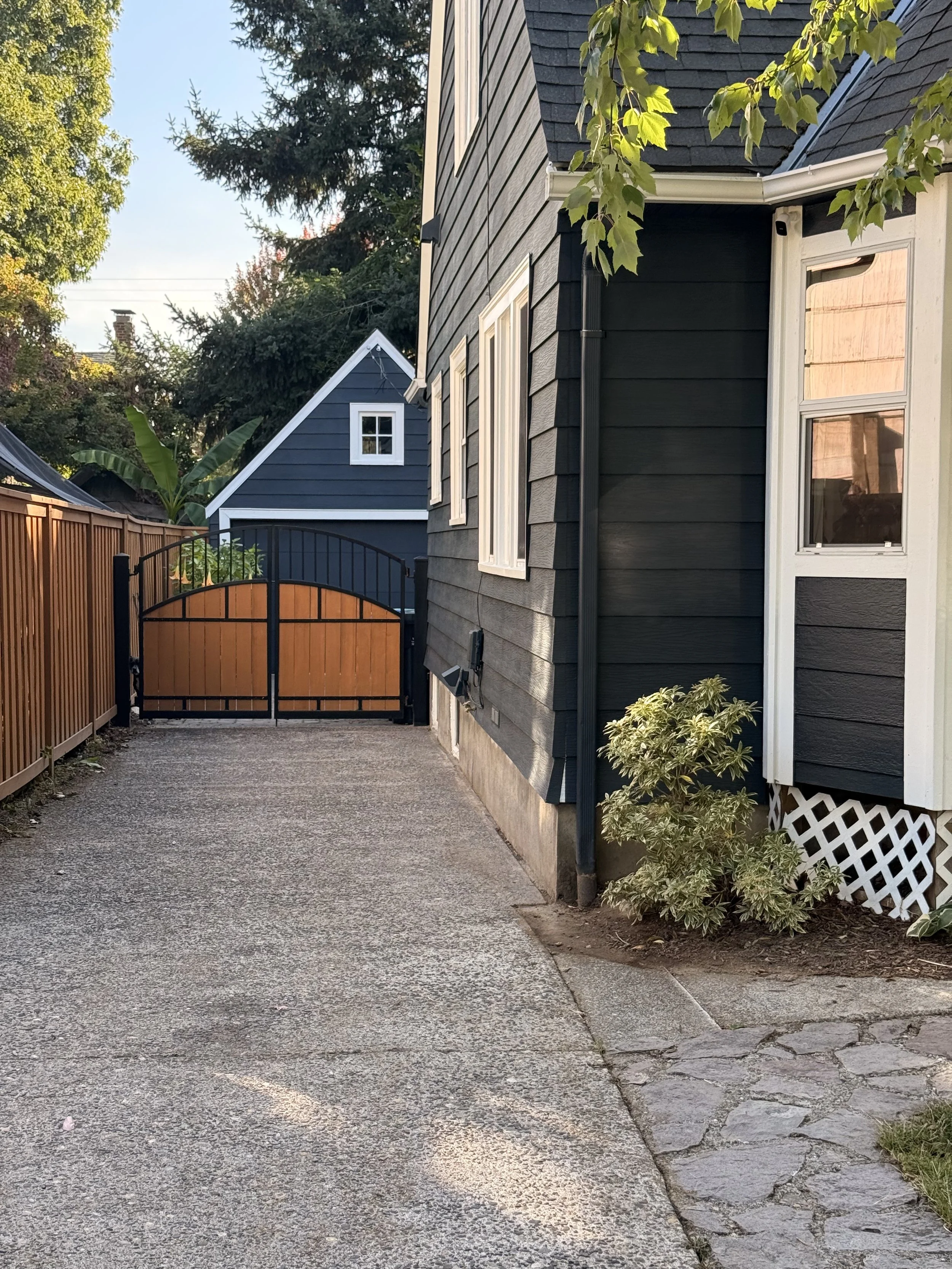 Residential backyard path with gravel surface, wooden fencing, a black gate, plants, and a house with dark siding and white window frames.