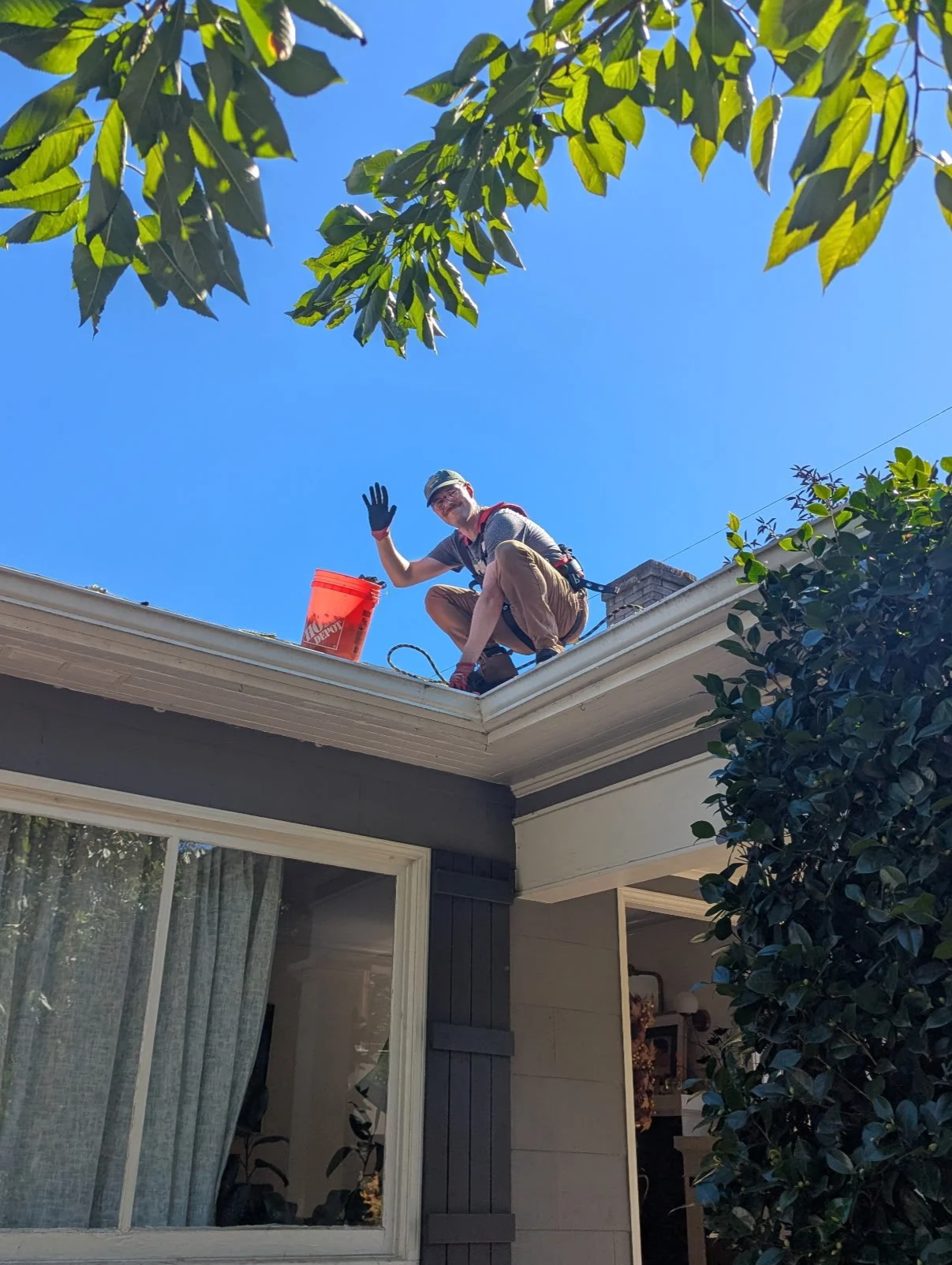 A person sitting on a roof edge waving, with a bucket beside them, looking down at the camera, under a clear blue sky, trees surrounding the house.