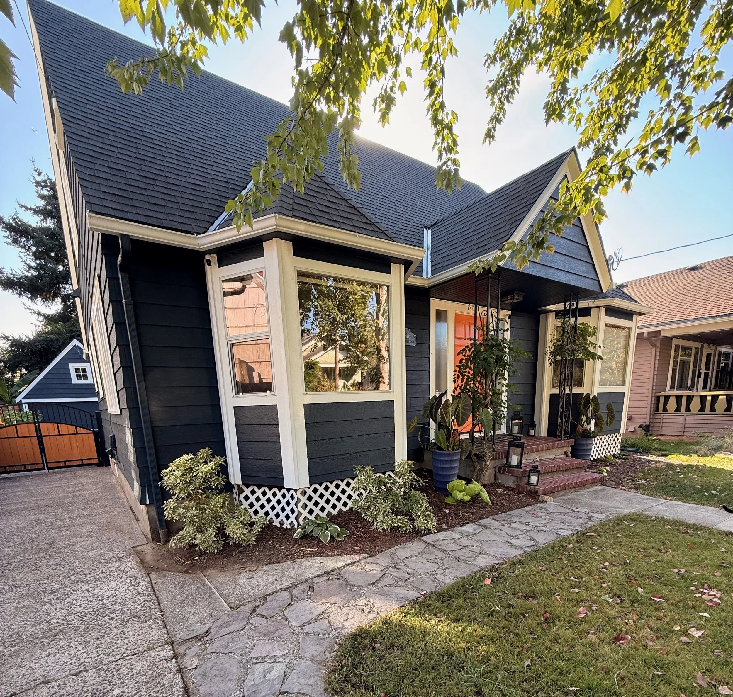 A house with dark blue shingles and white trim, with a large window and a small porch. The house is surrounded by a garden with plants and trees, and a brick walkway and a gravel driveway are visible. The sky is clear and sunny.