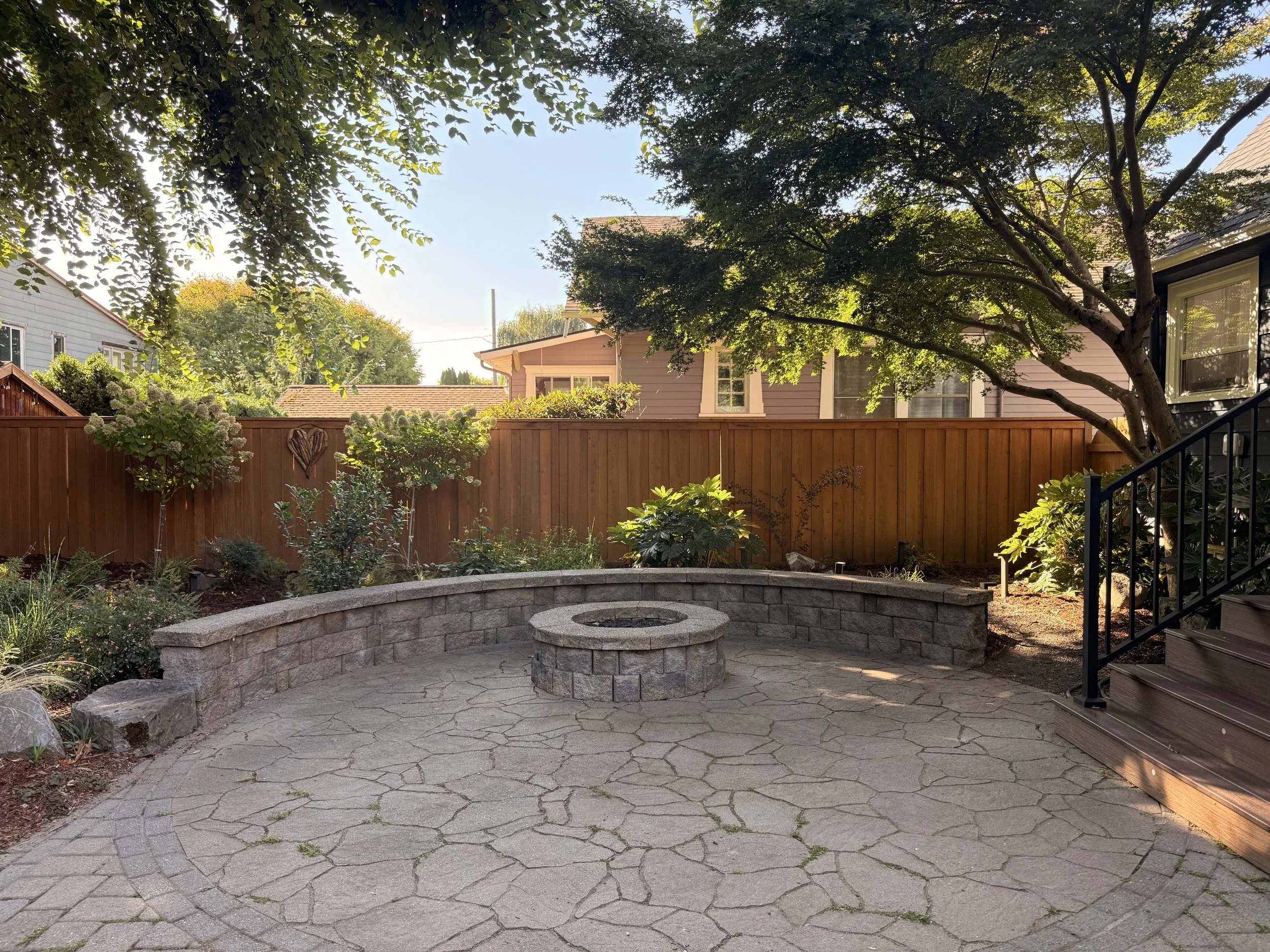 Backyard with stone patio, a fire pit, lush green plants, trees, a wooden fence, and residential houses with rooftops in the background, under a clear blue sky.