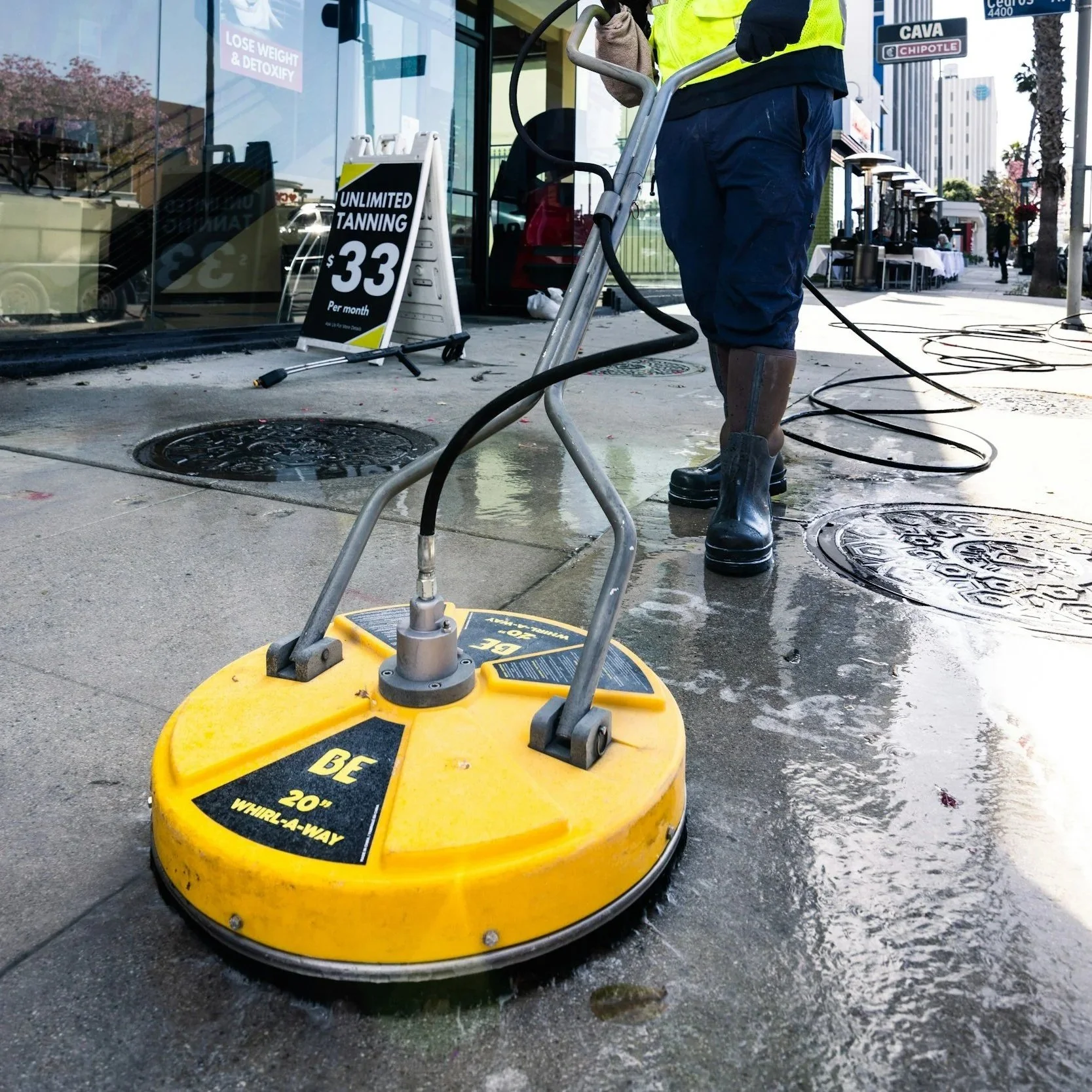A worker using a large yellow industrial scrubber while cleaning the street in front of a storefront with a sign advertising unlimited tanning for $33 per month.