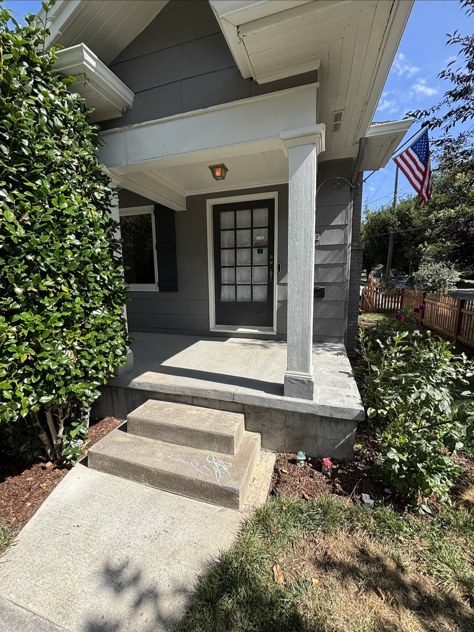 Front porch of a modern gray house with a black door, white trim, and a small front garden with green shrubs. A clean concrete walkway leads to the steps of the porch, and an American flag is flying in the yard.
