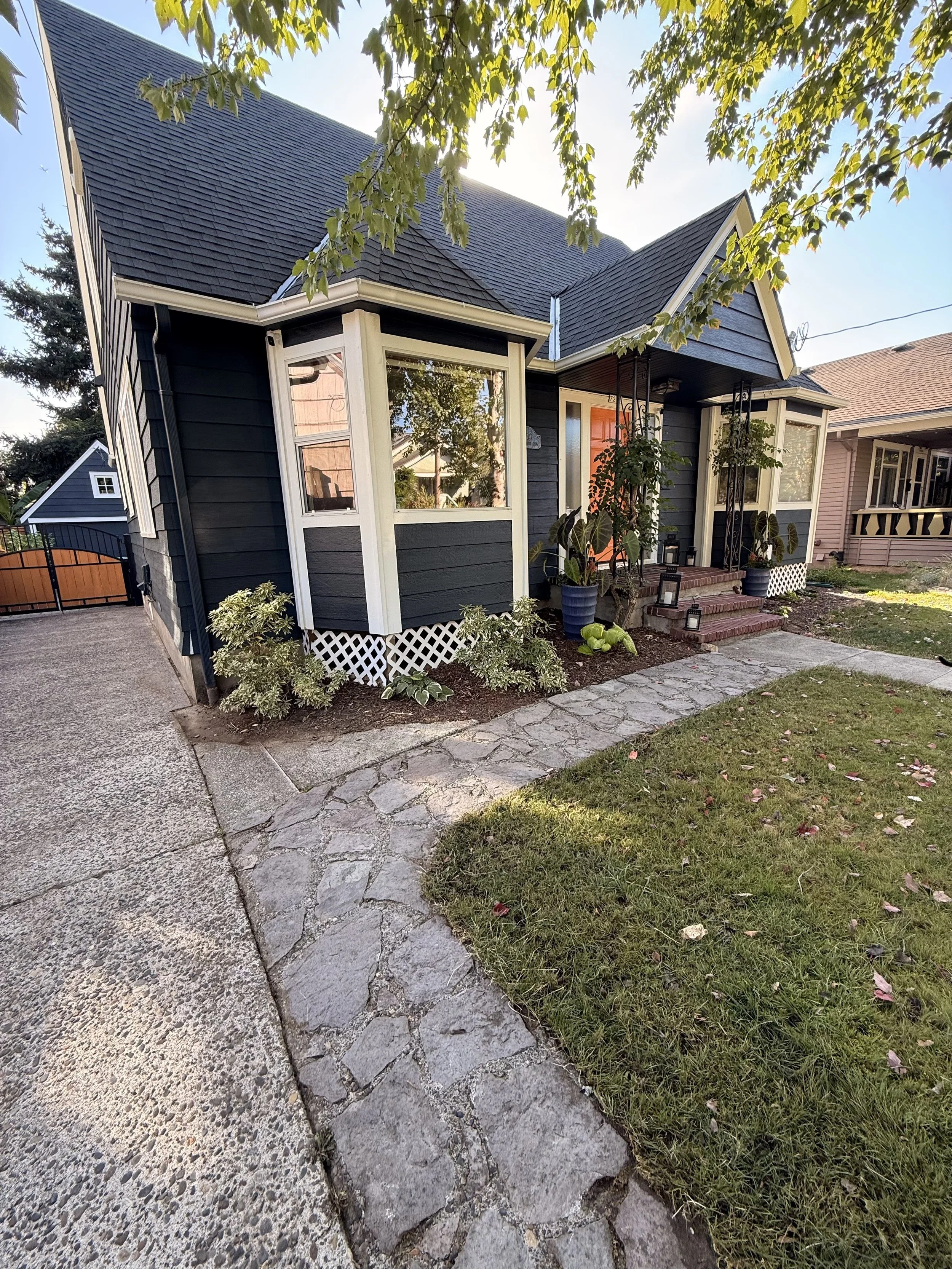 A house with black siding and white trim, front porch with steps, potted plants, and lanterns, surrounded by a lawn and a sidewalk, with trees and neighboring houses in the background.