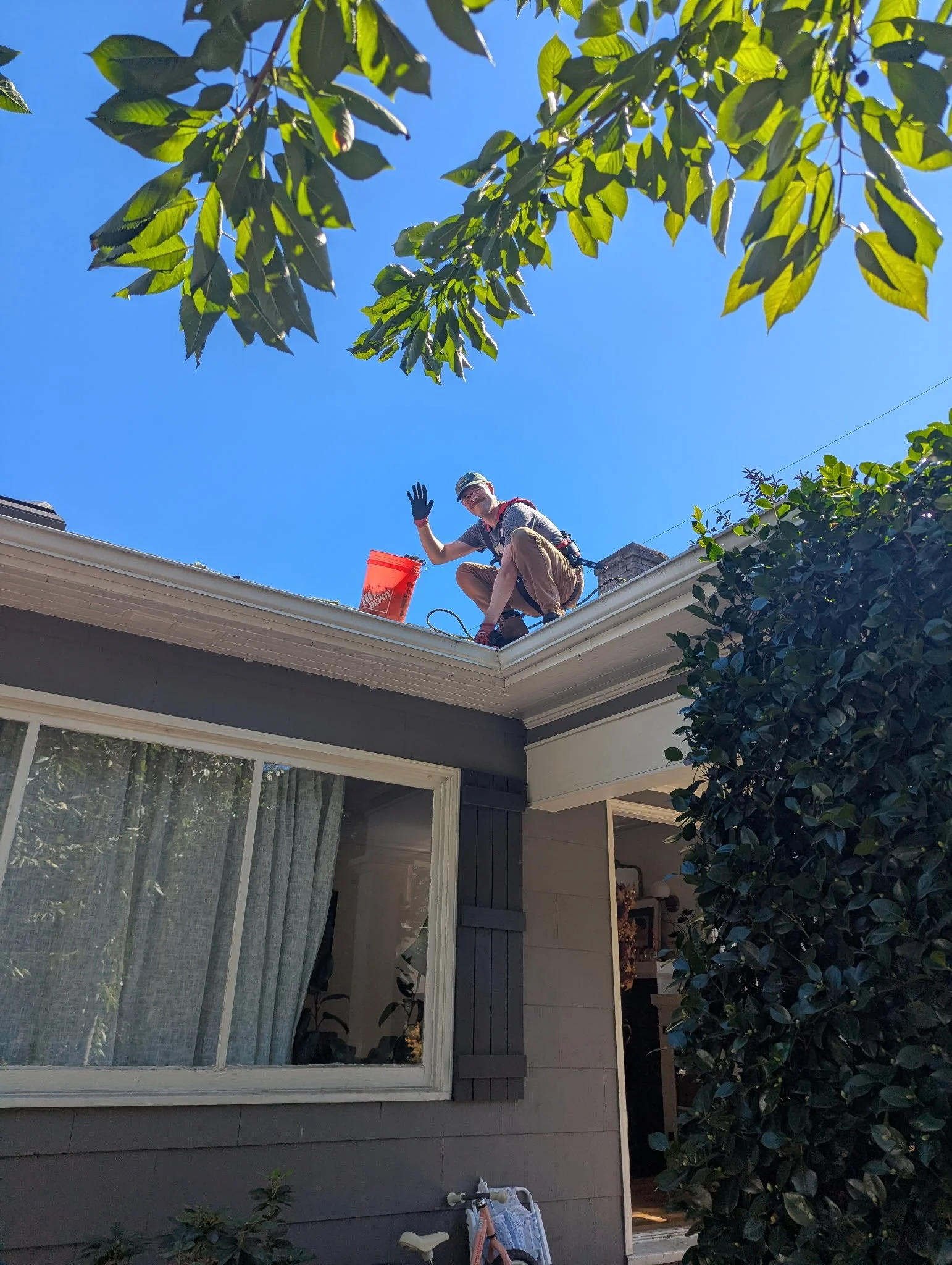 A person wearing glasses, a hat, and gloves on a roof, waving. They have an orange bucket beside them, and a harness is visible. The house has siding and large windows with curtains, surrounded by trees and bushes under a clear blue sky.