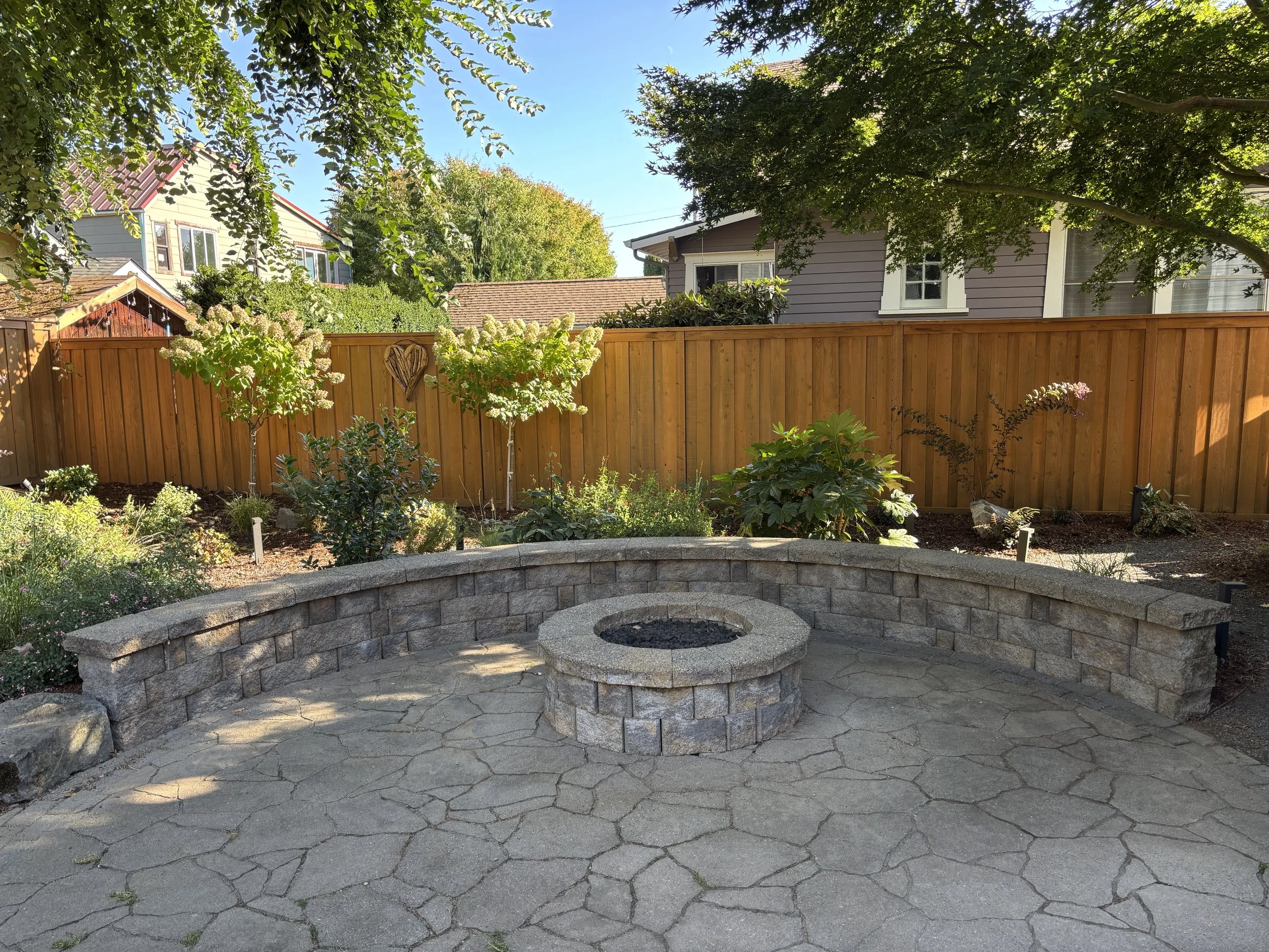 A backyard with a circular stone fire pit and a surrounding stone patio. A wooden fence runs along the back, with green trees and bushes, and neighboring houses visible beyond. Sunny weather with a clear blue sky.