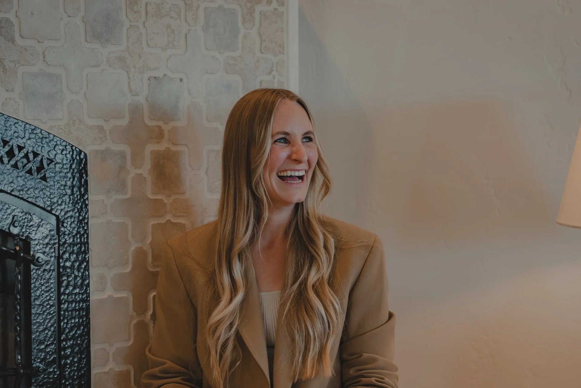 A woman with long wavy blonde hair, wearing a brown blazer, smiling and looking to her right, sitting indoors near a textured wall and a black metal decorative element.
