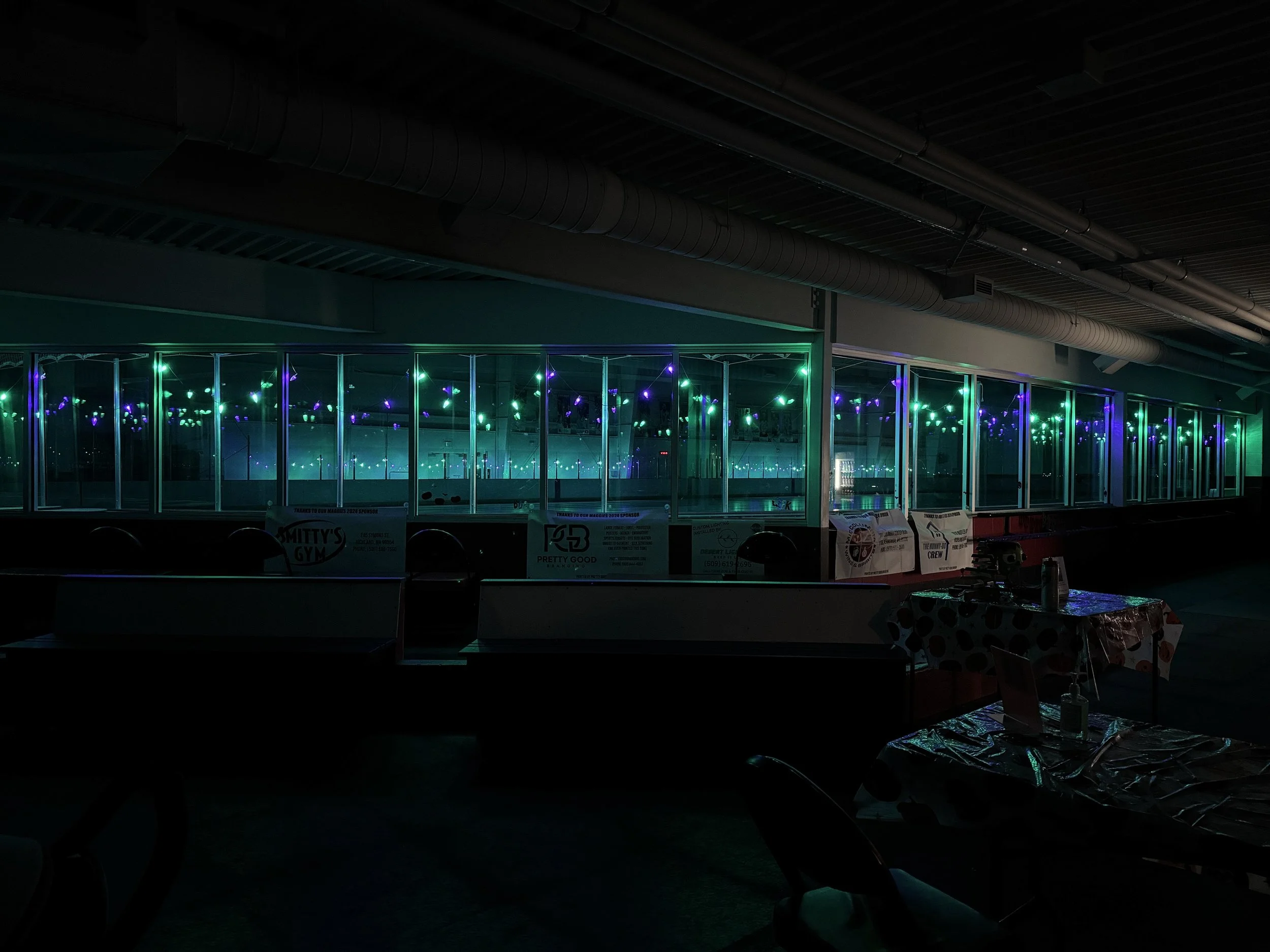 A dimly lit indoor space with illuminated reflective surfaces and faint purple and green decorative lights, possibly an art installation or nightclub interior.