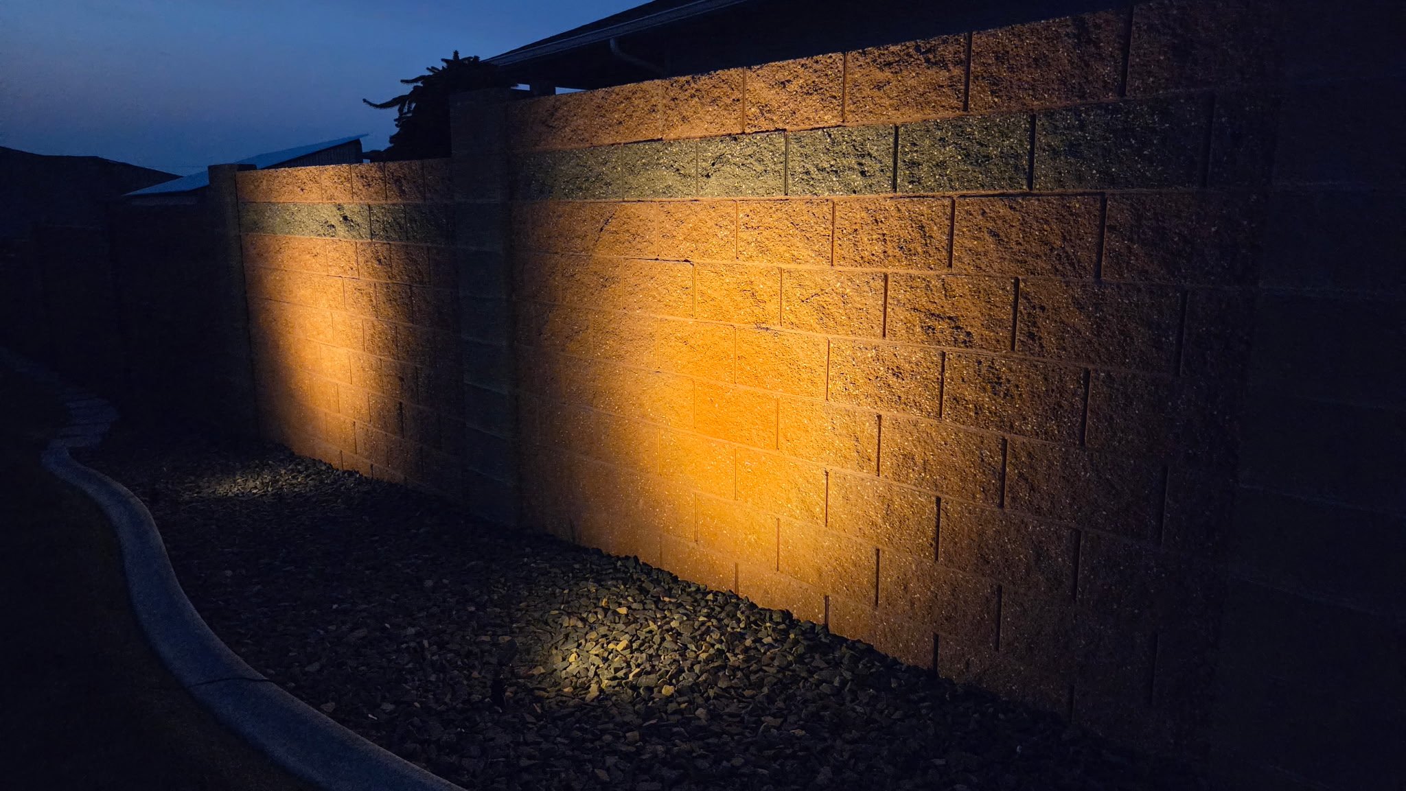 A brick privacy wall illuminated by outdoor lighting at dusk, with a cloudy sky in the background.