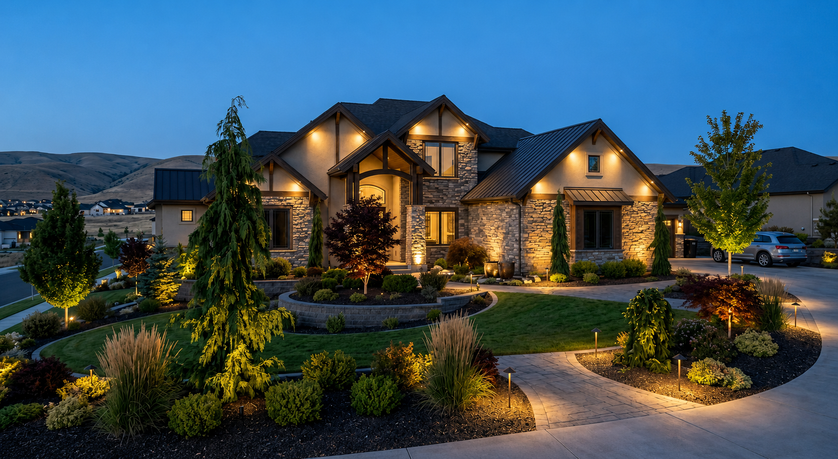 Night view of a well-lit, modern suburban house with a landscaped front yard and driveway, surrounded by hills.