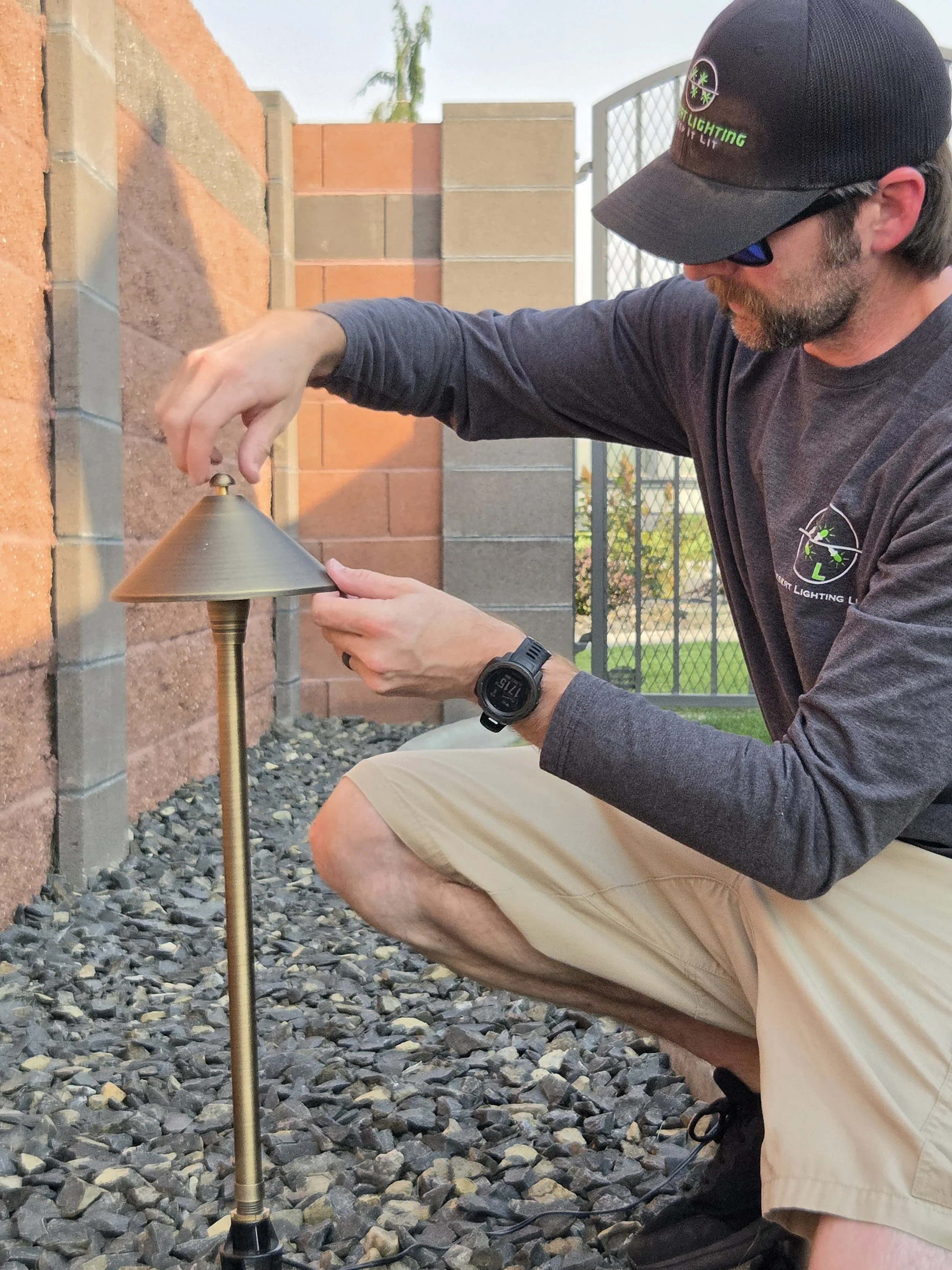 A man wearing a baseball cap and sunglasses is installing or adjusting a landscape lamp on a gravel pathway next to a brick wall, with a fence and greenery in the background.