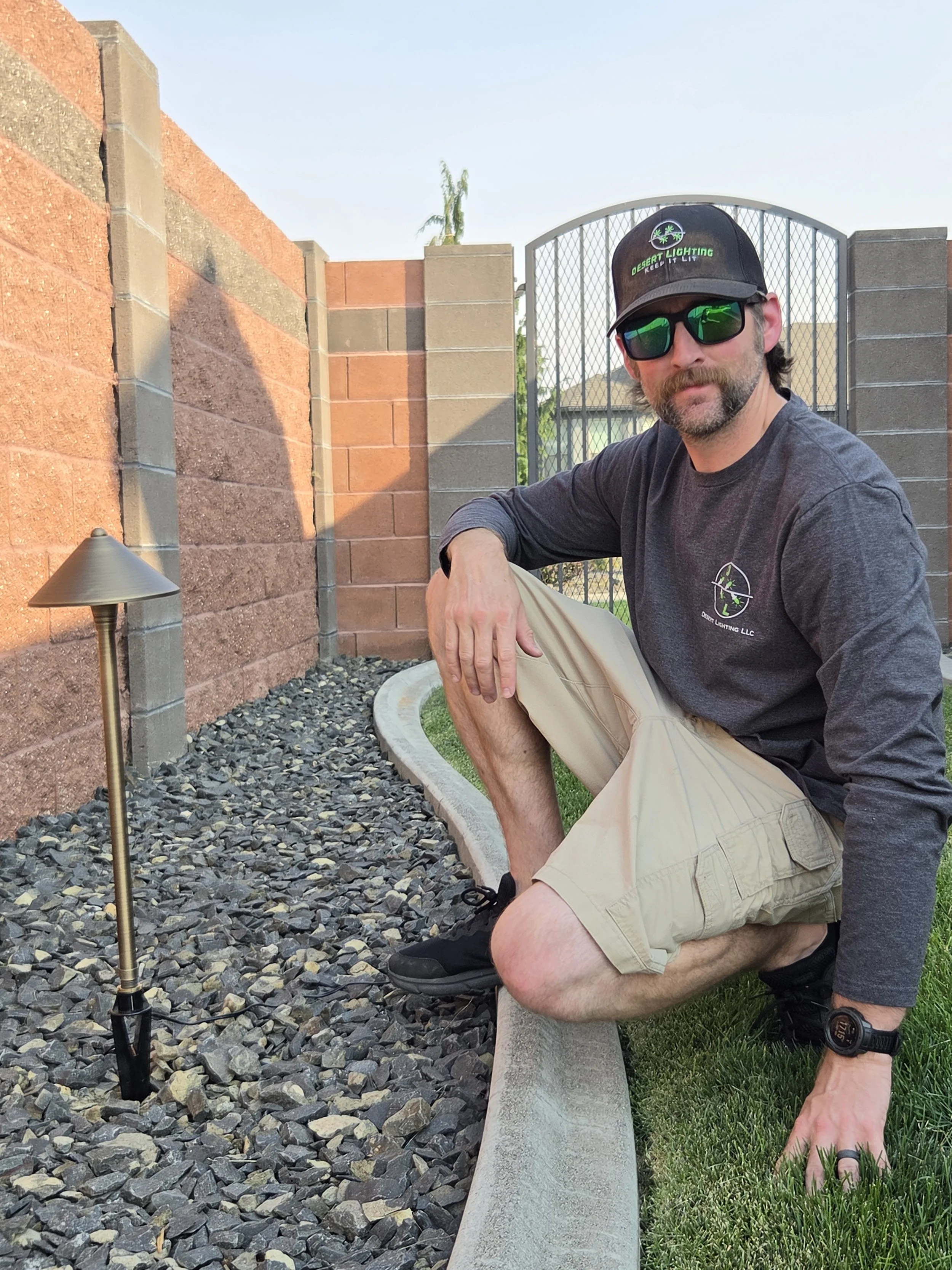 A man with a beard wearing sunglasses, a black cap, a gray long sleeve shirt, and khaki shorts is kneeling next to a concrete bench in a backyard. The backyard has a red brick wall, a metal gate, and green grass. There is a small outdoor lamp post and a gravel area next to the bench.