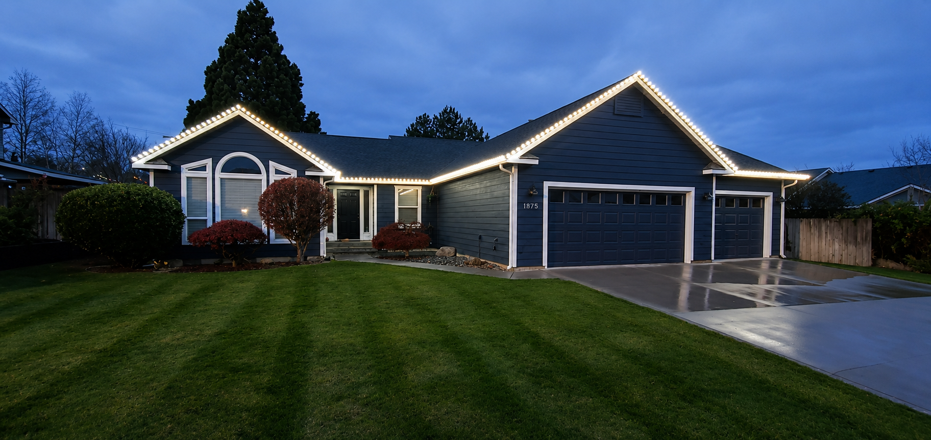 A blue house with a dark roof outlined with white holiday lights, with a well-maintained lawn and a wet driveway, during the evening.