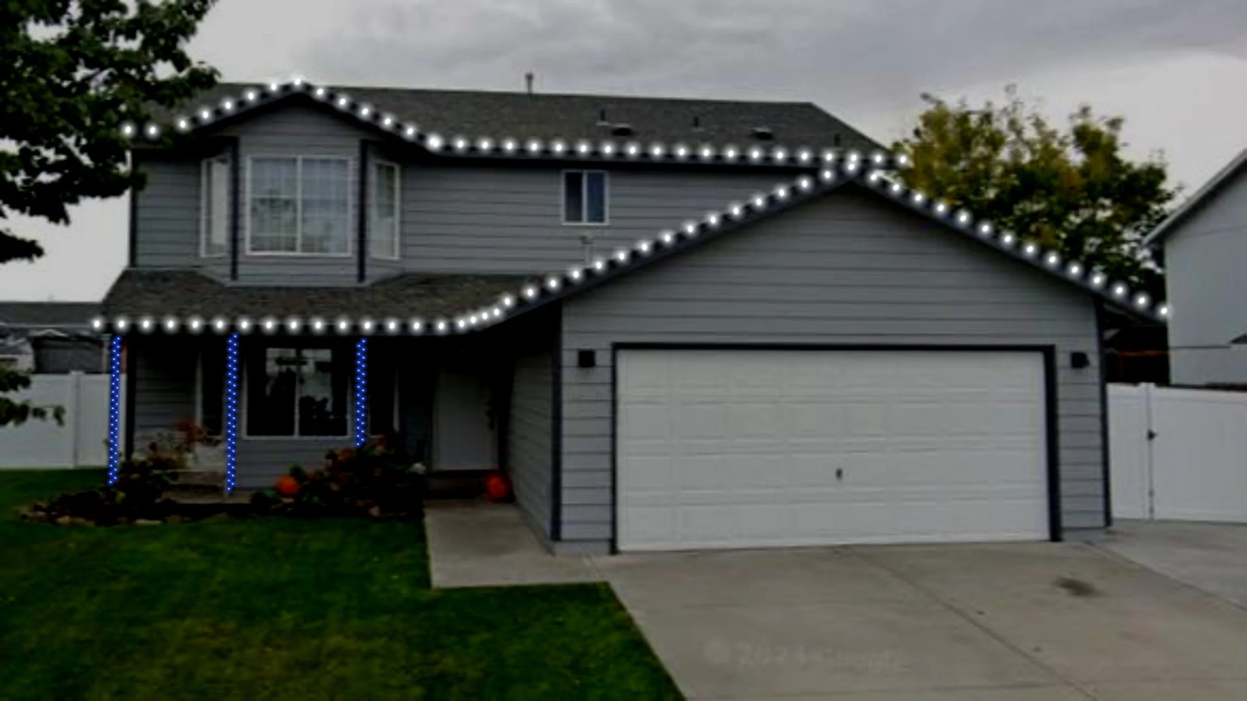A two-story house decorated with string lights along the roofline and porch columns, with a well-maintained front yard, a concrete driveway, and a white garage door. There are trees and neighboring houses visible in the background.