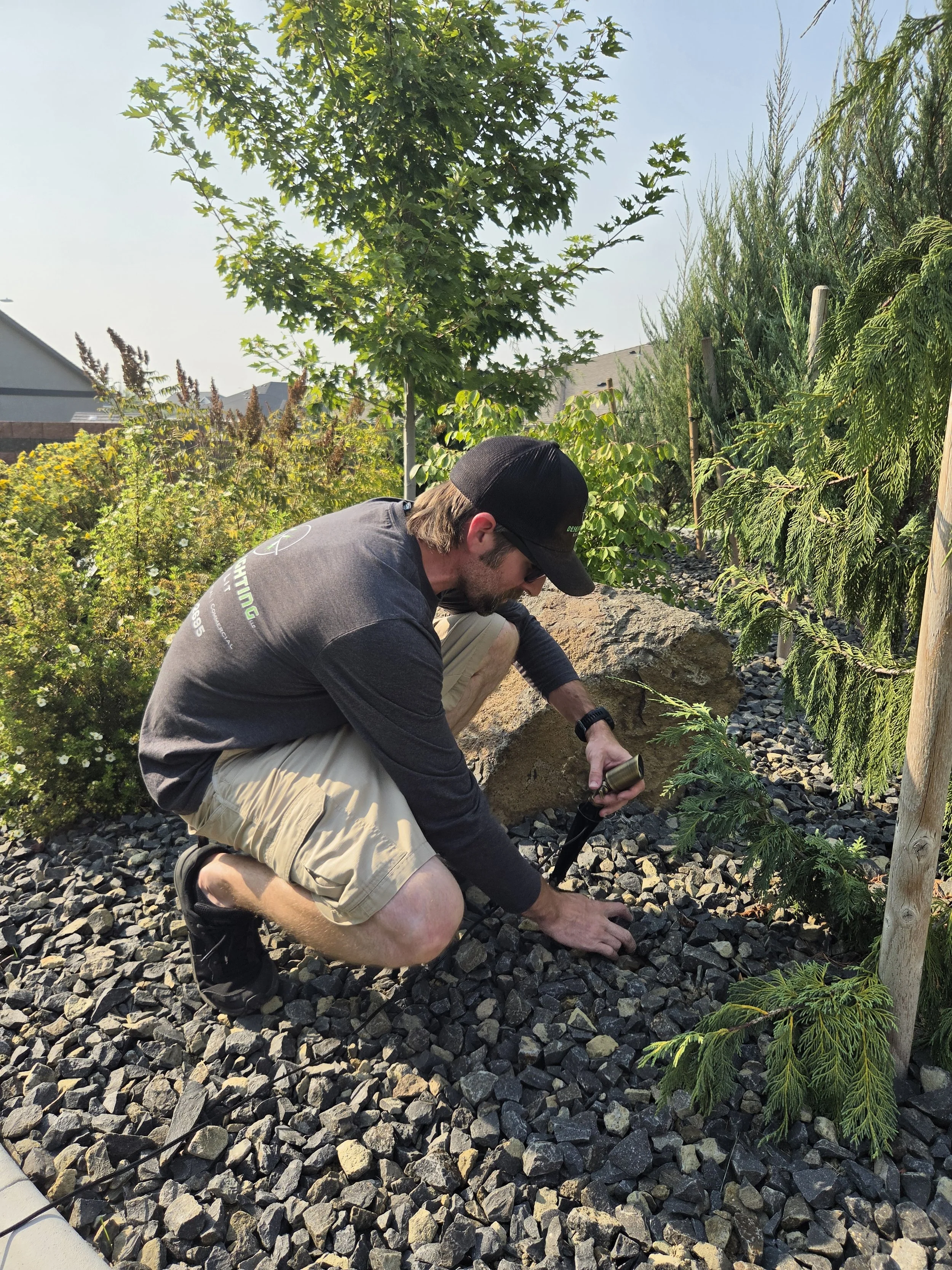 A man wearing a cap, sunglasses, and casual outdoor clothing kneels on a rocky ground with small gravel, working with a drill or similar tool near a landscaped garden area with various green plants and a young tree. Residential houses and a fence are