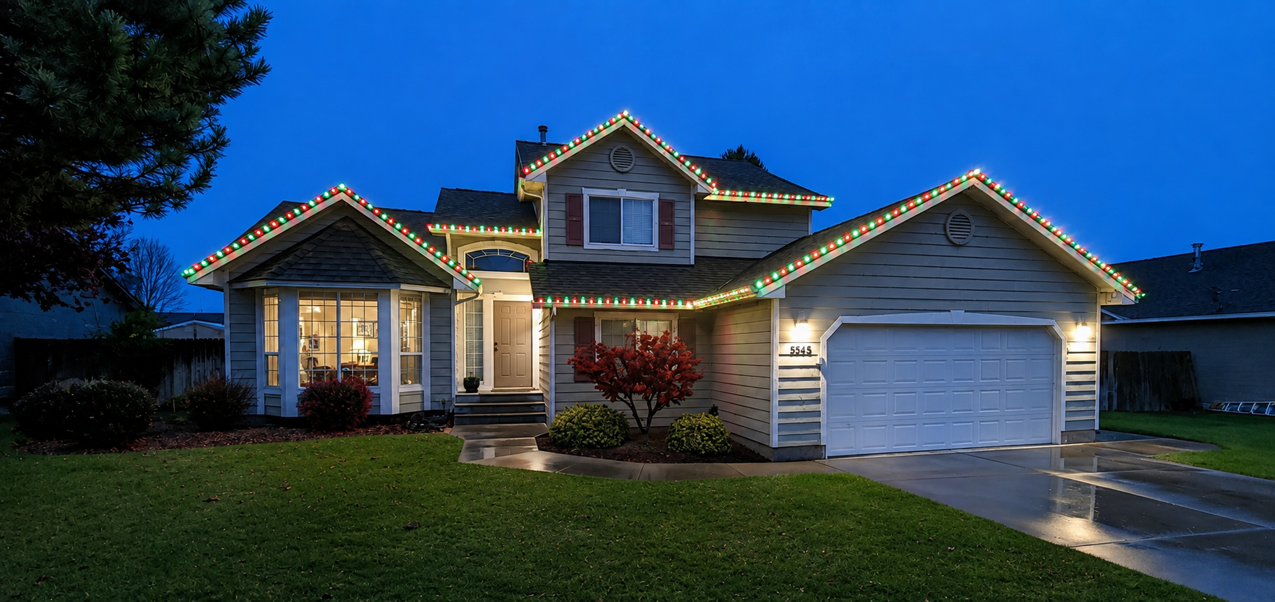 A suburban house decorated with red and green Christmas lights along the roofline during dusk. The house features a bay window on the left, a front door with steps, a driveway on the right, and well-maintained front yard with shrubs and a small tree.