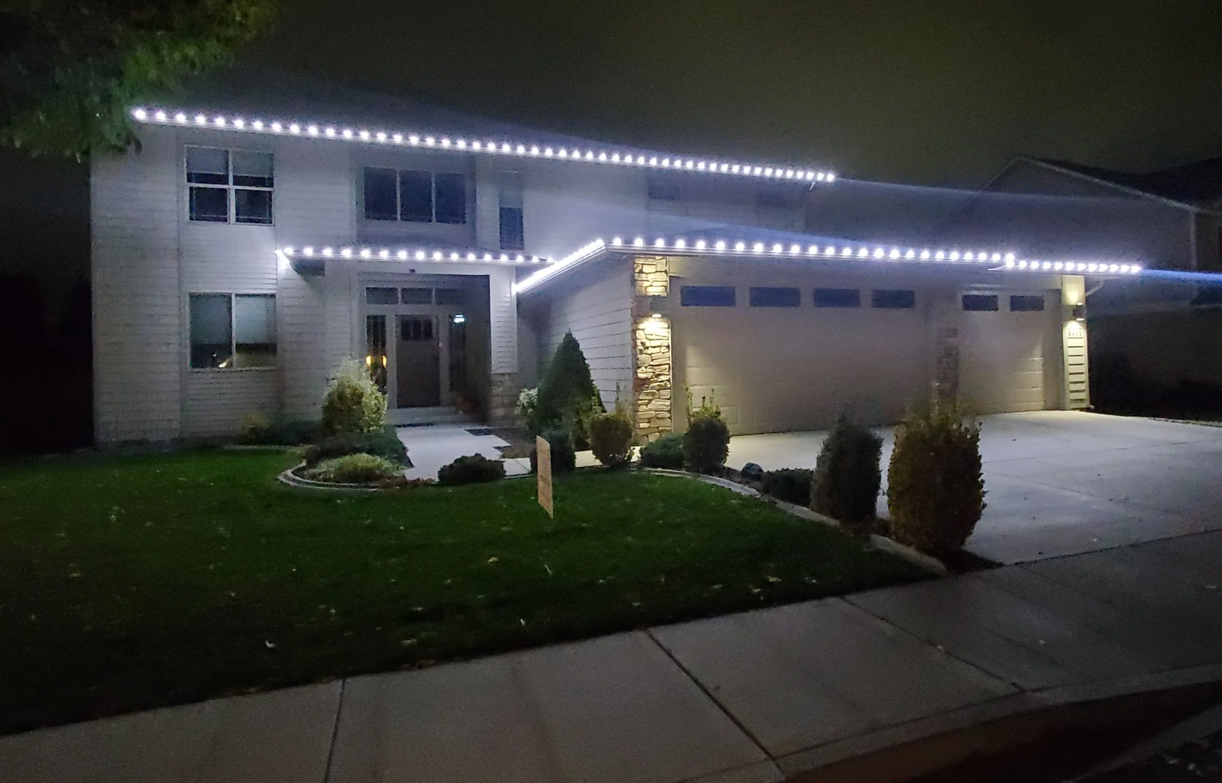 Nighttime photo of a house decorated with white string lights along the roofline and garage, with a landscaped yard and sidewalk in the foreground.