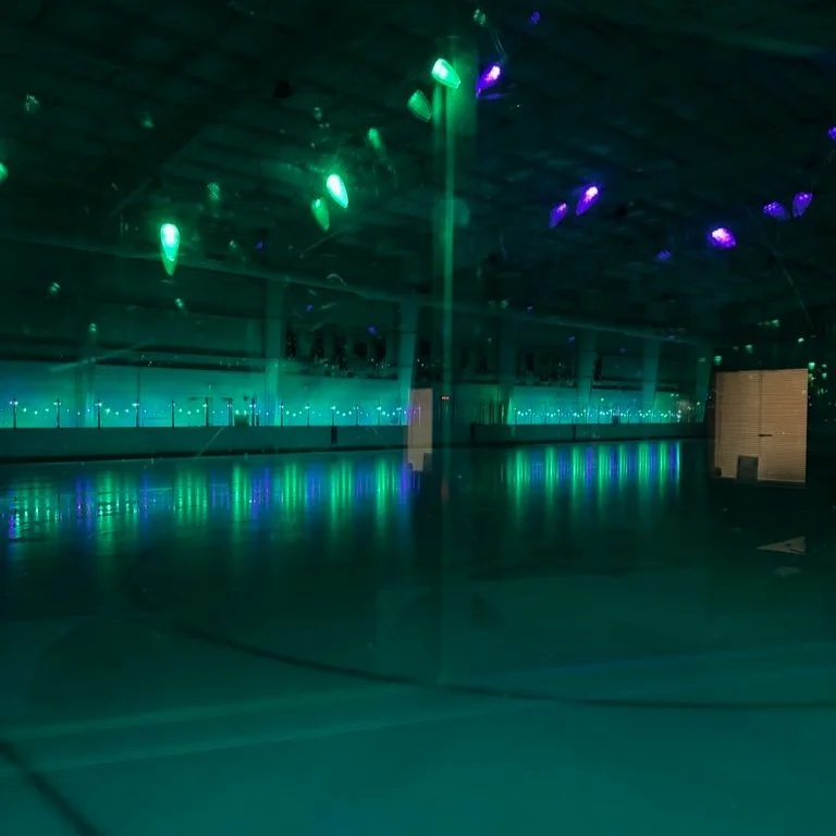 An empty indoor ice skating rink illuminated by blue and green lights with reflections on the ice.