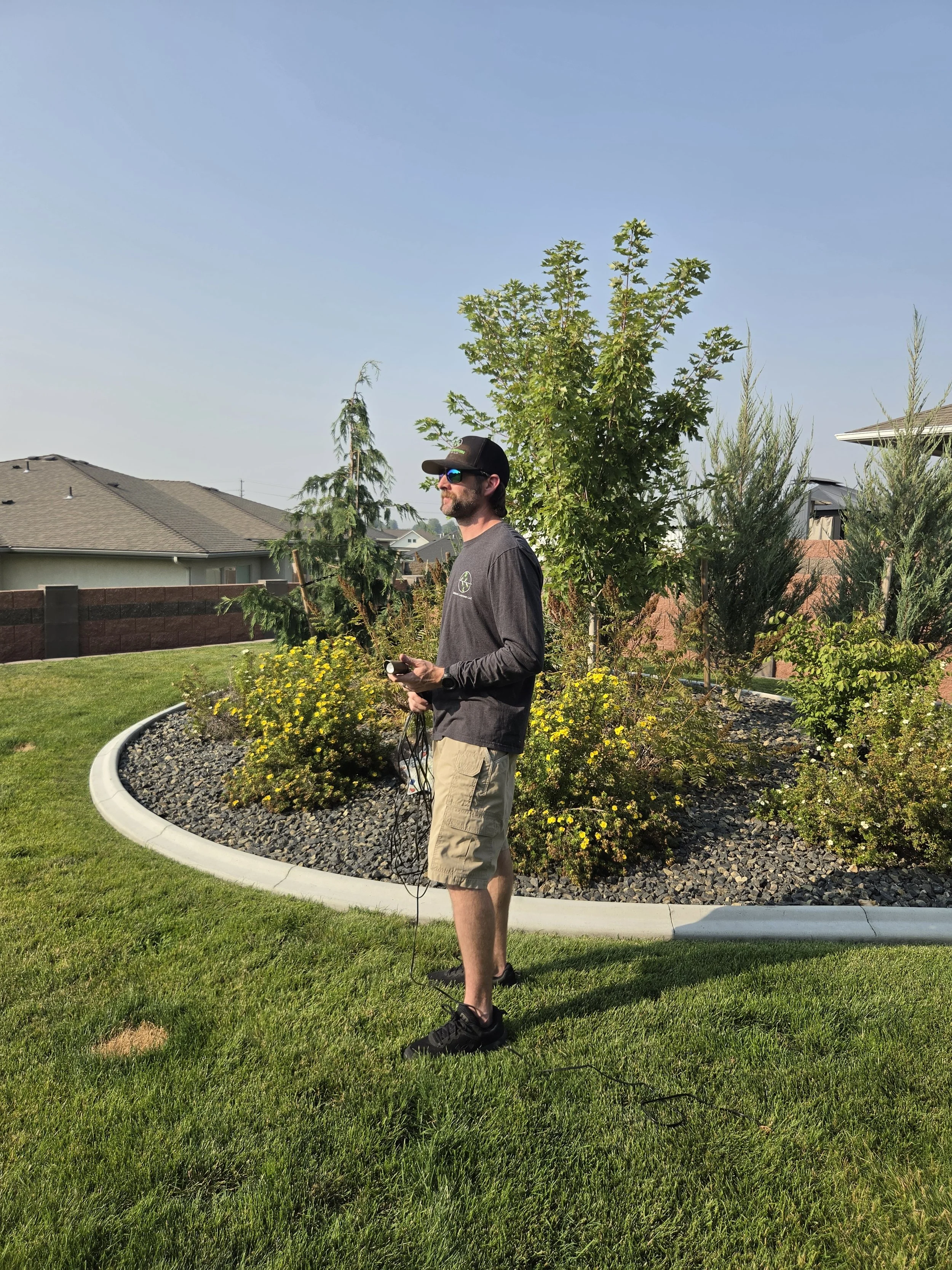 Man with a beard, wearing sunglasses, a cap, a gray long-sleeve shirt, and khaki shorts stands in a backyard with landscaped garden, holding a remote control. Residential houses and trees are in the background, under a clear blue sky.
