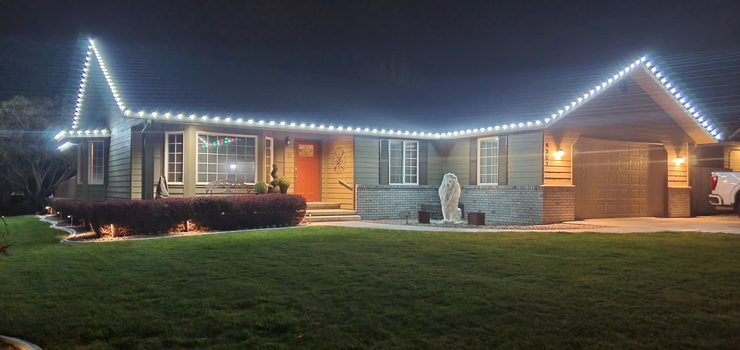 A house at night decorated with white holiday lights along the roofline, with a well-maintained front yard, shrubs, and a lawn, featuring a lion sculpture near the front steps and a car in the driveway.