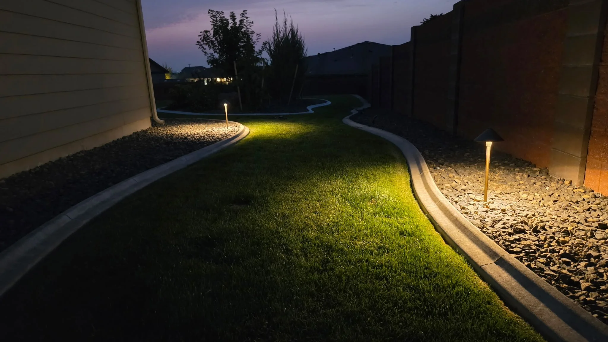 A backyard at dusk with a curved pathway lit by ground lights, surrounded by green grass and decorative rocks, enclosed by a fence, with trees and houses visible in the background.