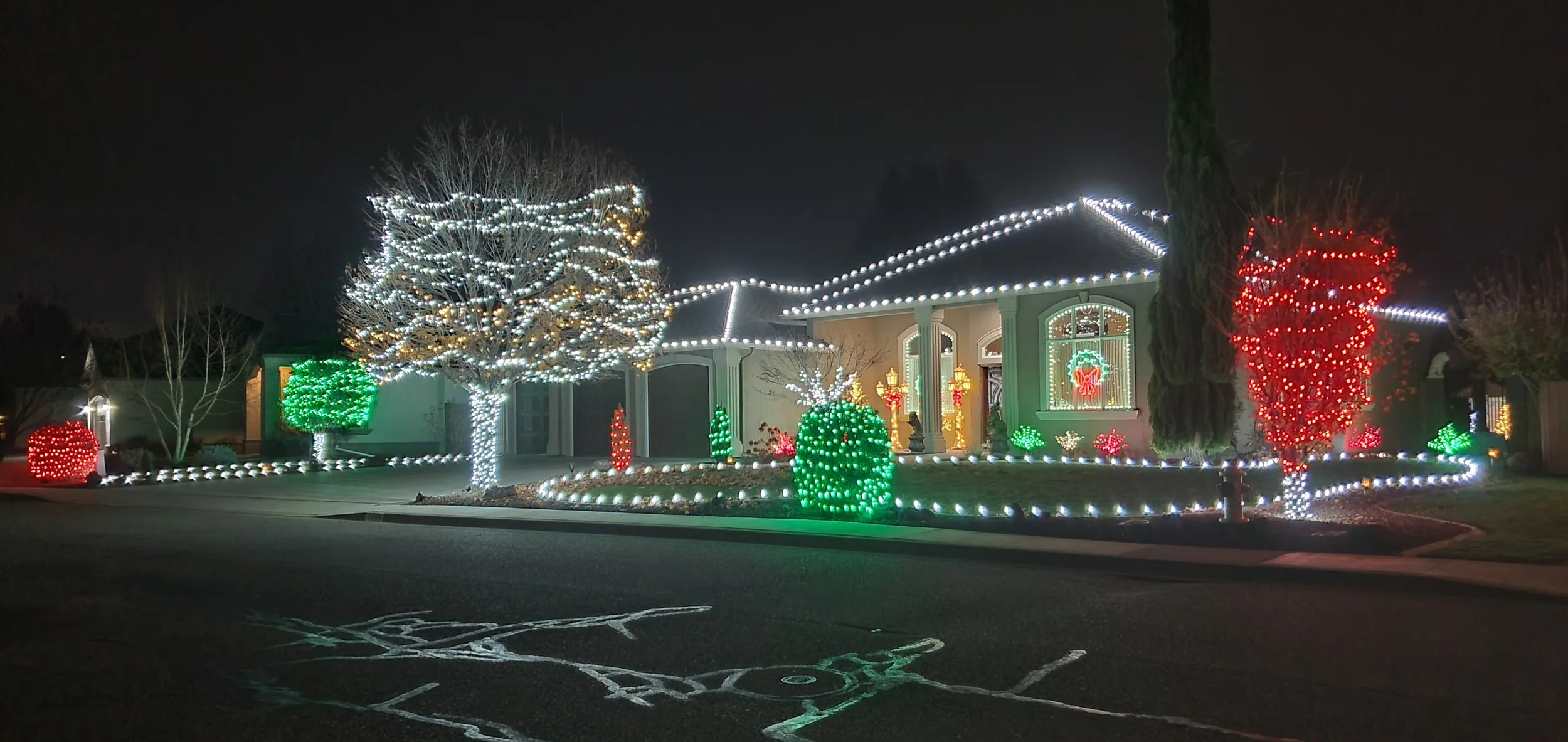 A house decorated with Christmas lights at night, including illuminated trees, bushes, and holiday-themed window displays.