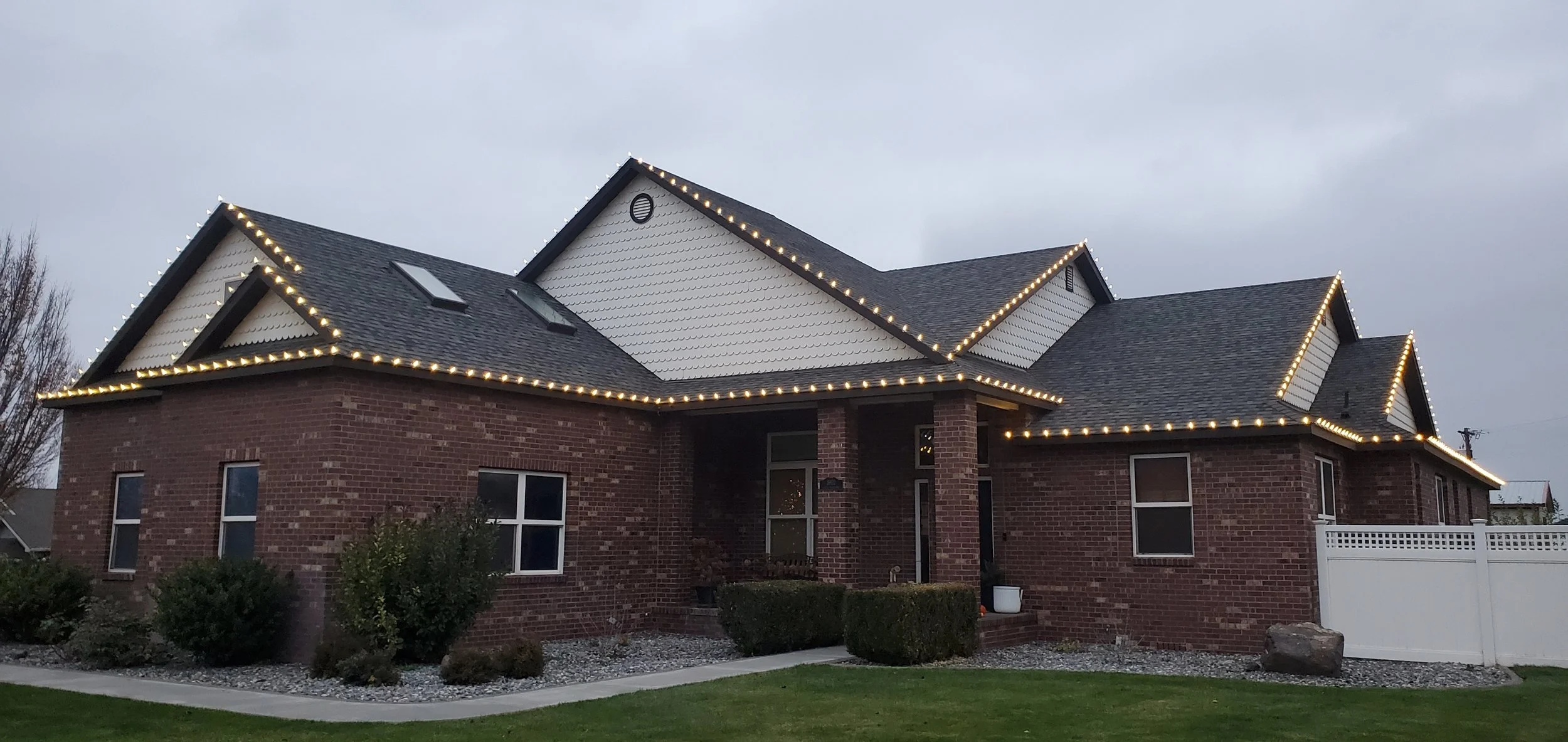 A two-story brick house decorated with white Christmas lights outlining the roofline, windows, and porch area, with a well-maintained lawn and bushes in front, under an overcast sky.