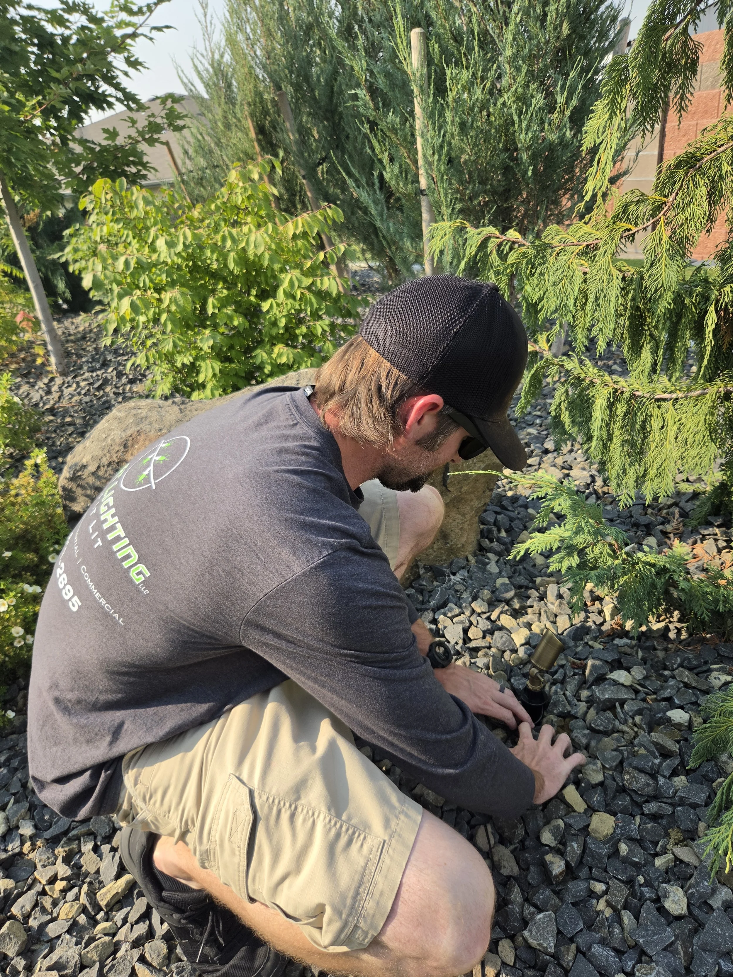 A man wearing a black cap, sunglasses, a gray T-shirt, and beige shorts kneeling on a gravel surface outdoors, working with a tool near evergreen trees and plants.