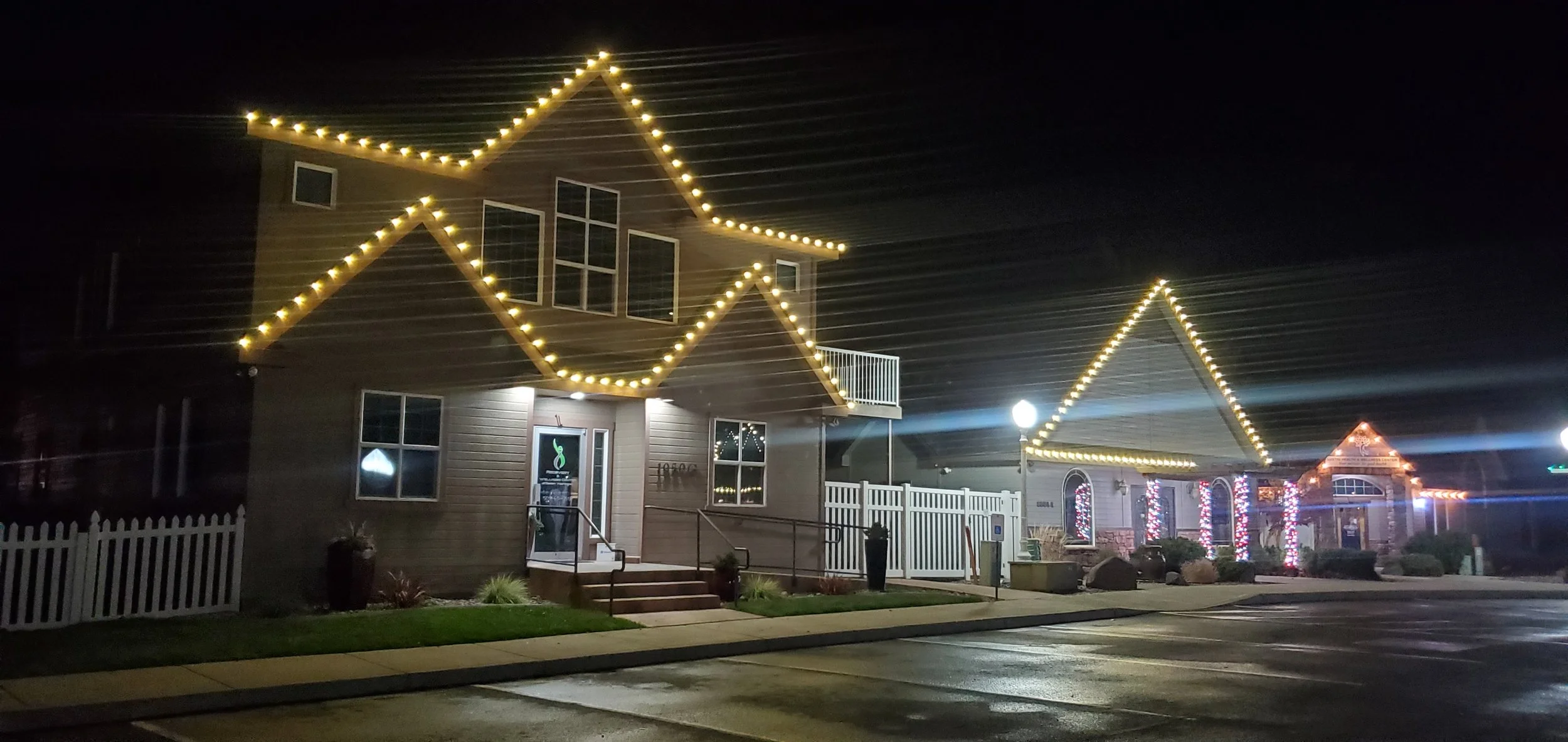 Night view of a building decorated with string lights outlining the roof and windows, with some Christmas lights visible on other buildings in the background.