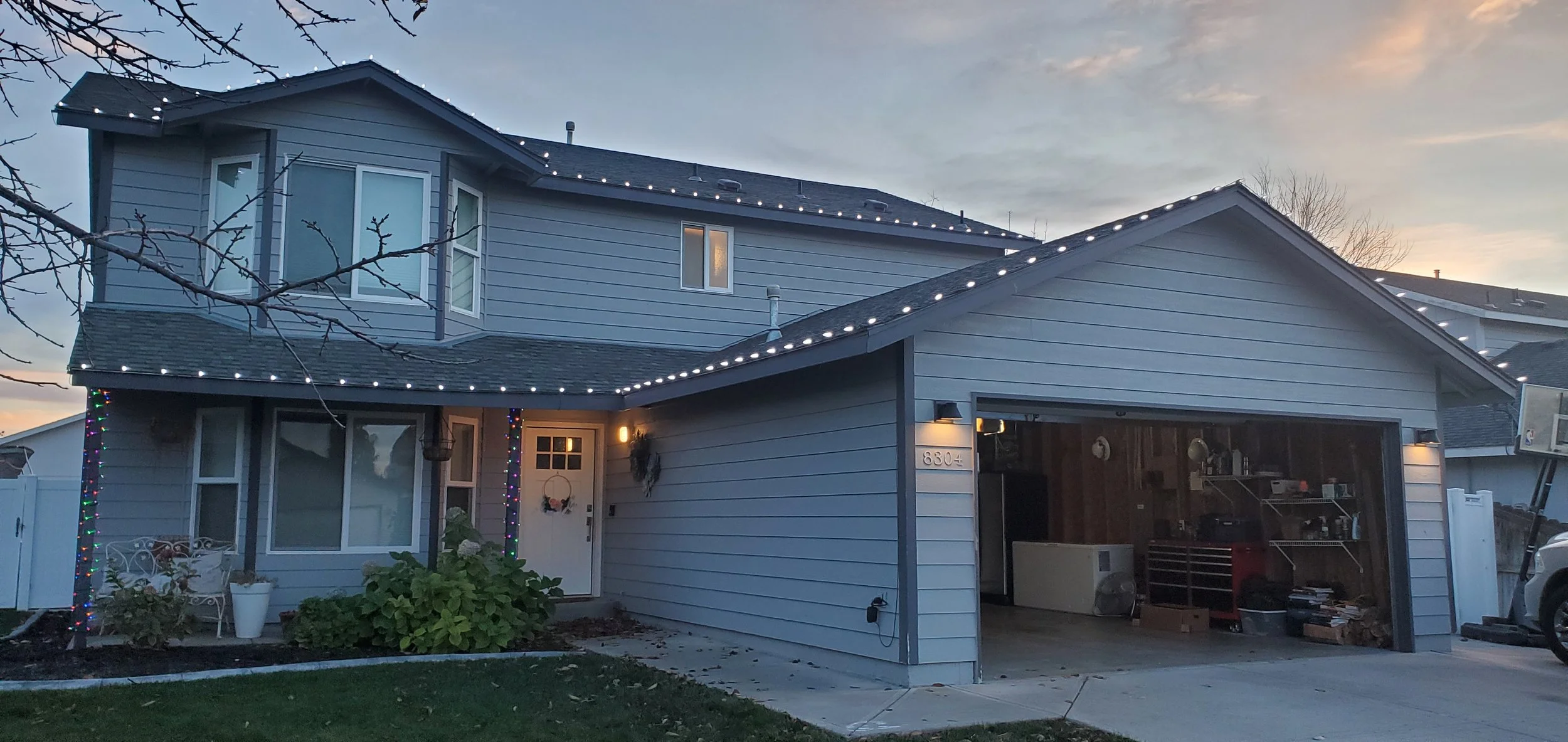 A two-story house decorated with white Christmas lights along the roofline, with some additional holiday lights on a pole near the front door. The house is light blue with white trim, and there's a garage with an open door showing items inside. The f