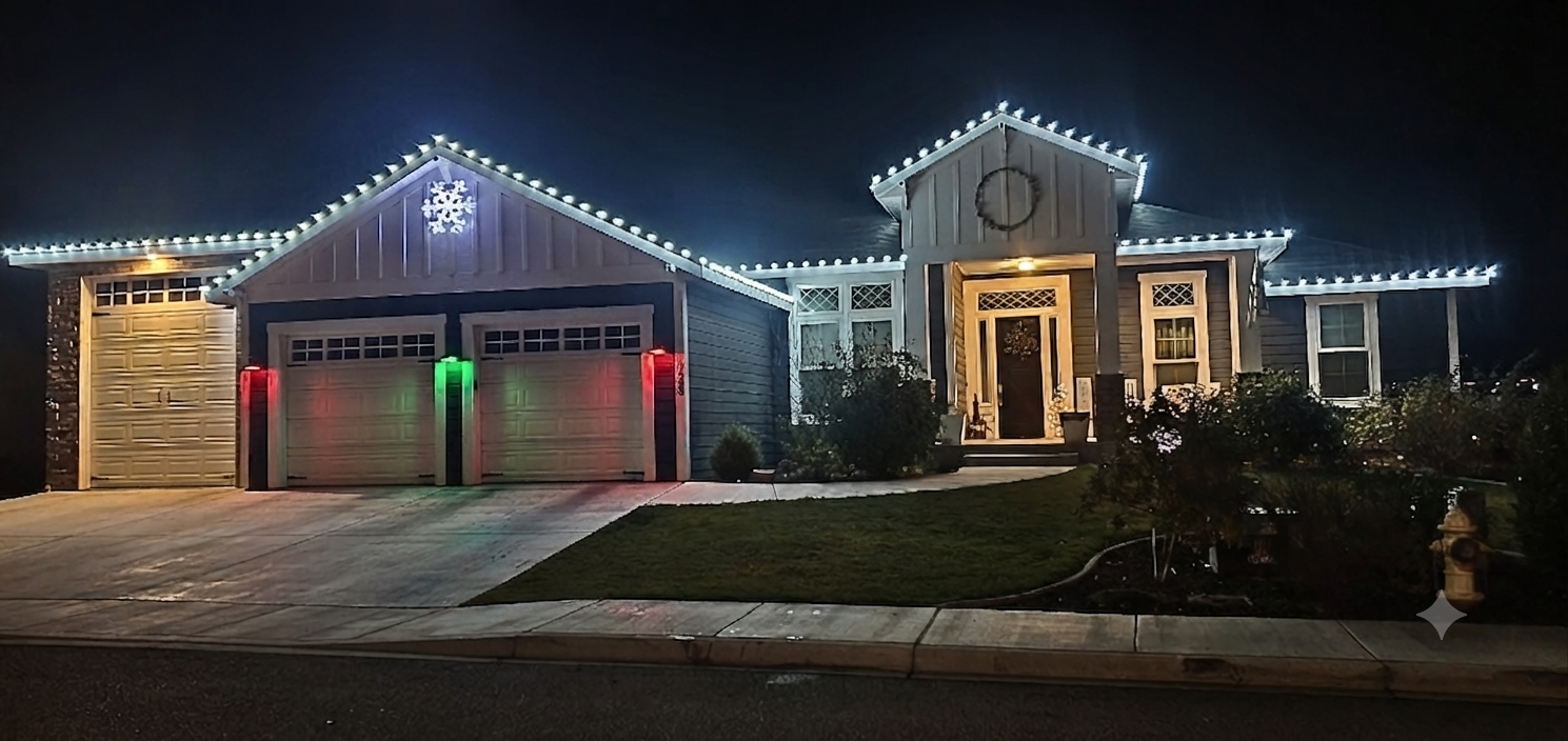 A house decorated with Christmas lights at night. The house has a two-car garage with lights on the roof edge, a snowflake decoration above the garage, and a wreath on the front door. The driveway and pathway are illuminated, with shrubs and a lawn i