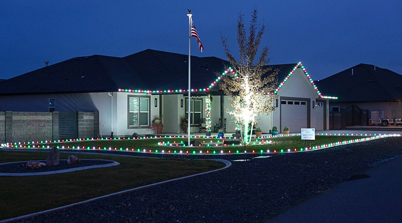 A house decorated with Christmas lights, including a Christmas tree illuminated with white lights in the front yard. American flag on a pole and a clear blue evening sky.