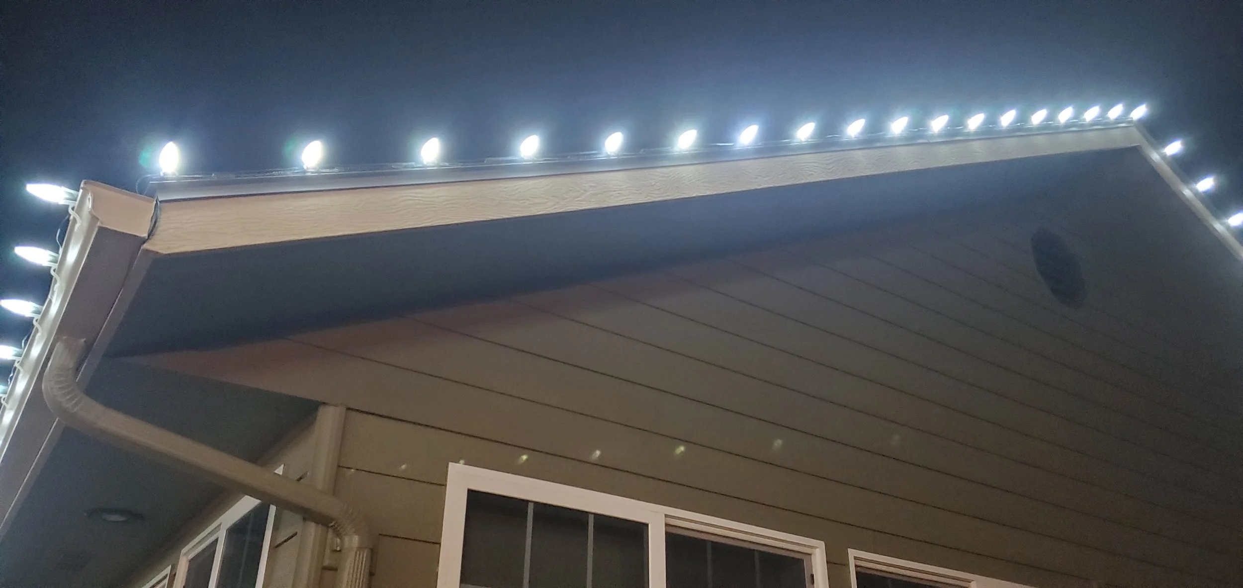 Nighttime view of house exterior with illuminated string lights along the roof edge, beige siding, and a window with a sliding door below.