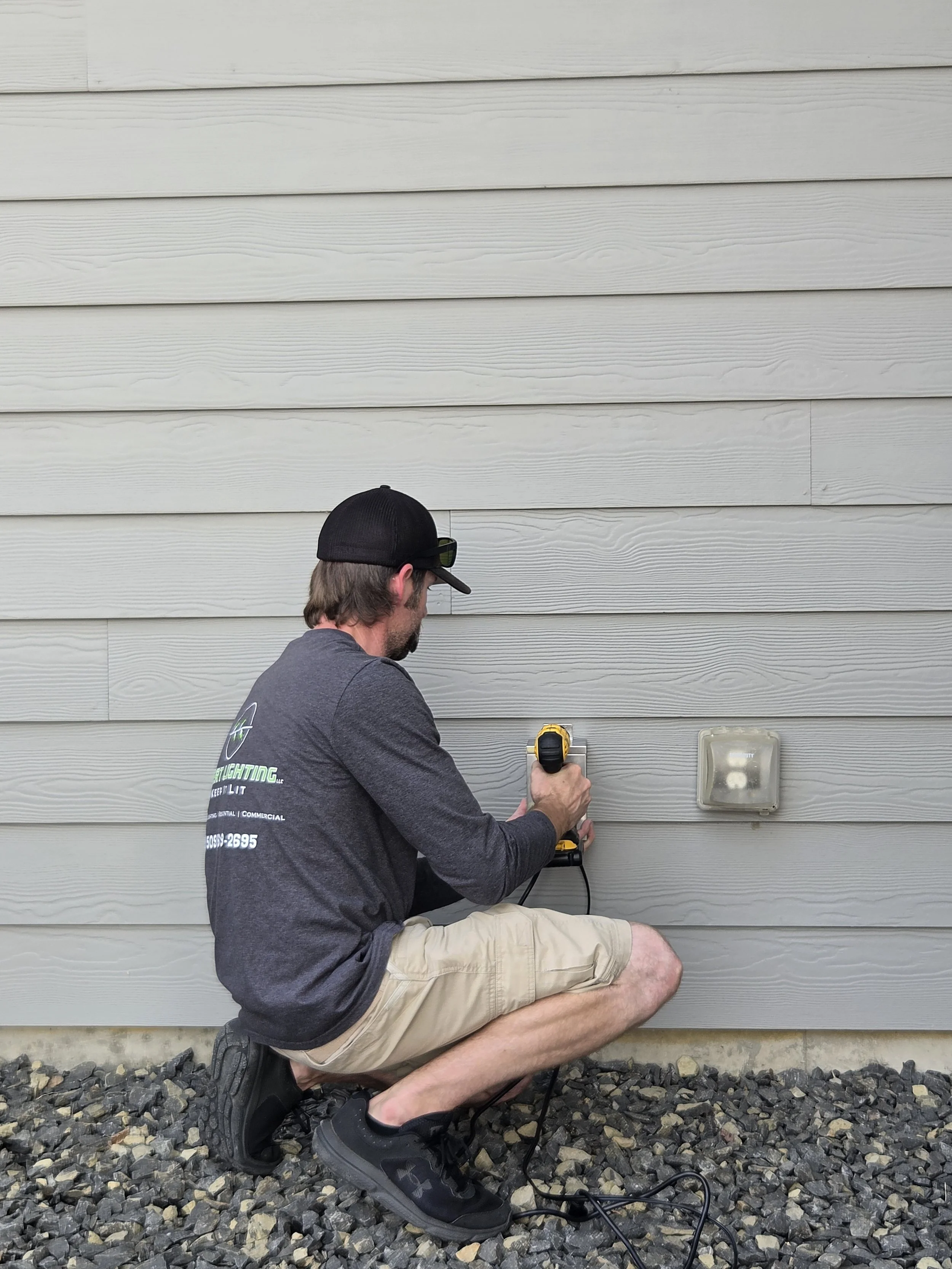 A man kneeling on gravel installing or repairing an outdoor wall-mounted light fixture with a cordless drill, wearing a black cap, glasses, a gray hoodie, and beige shorts.