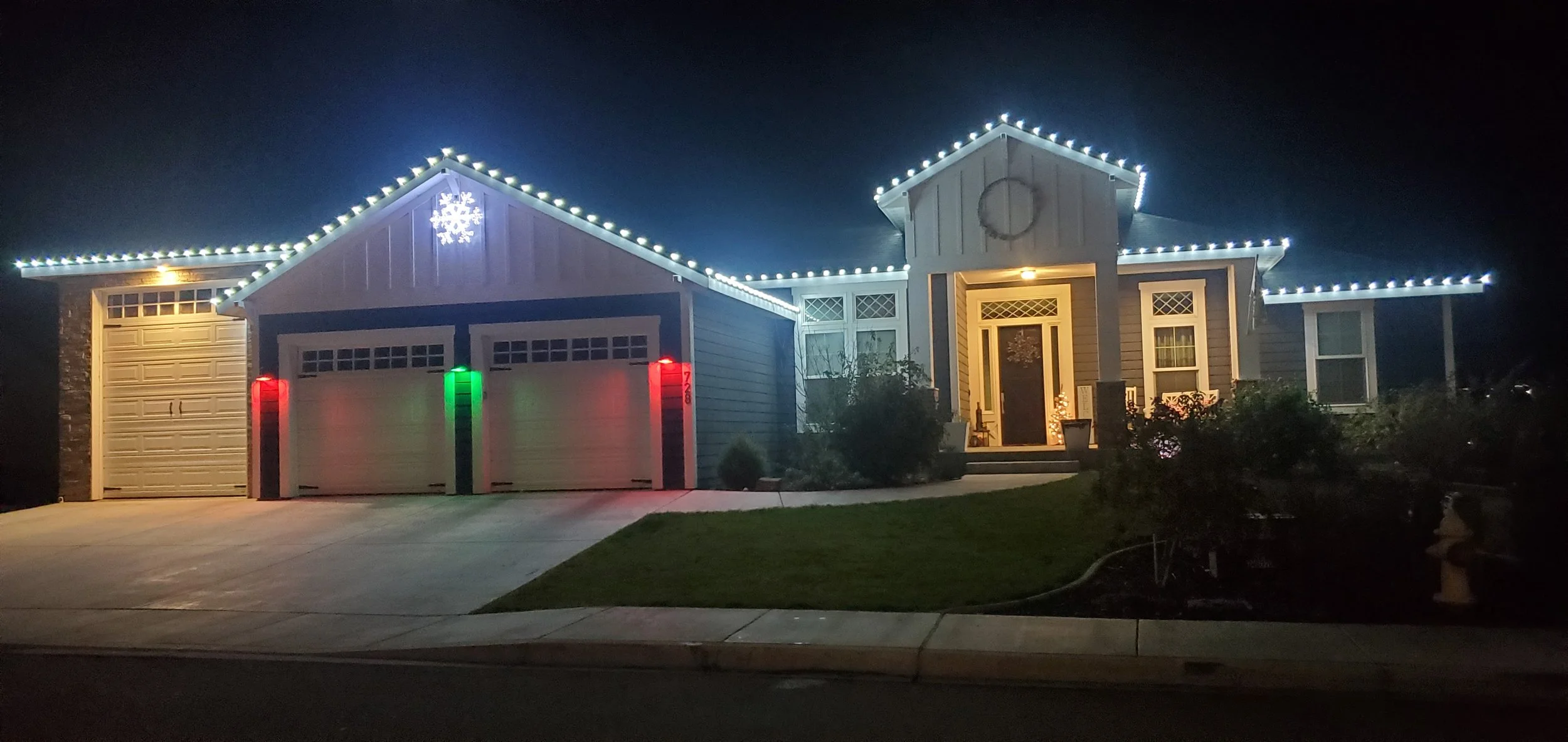 Nighttime exterior of a house decorated with white Christmas lights along the roofline and a snowflake decoration near the roof peak. The house has a two-car garage with a windowed garage door, and the driveway has a phone or alarm system intercom wi