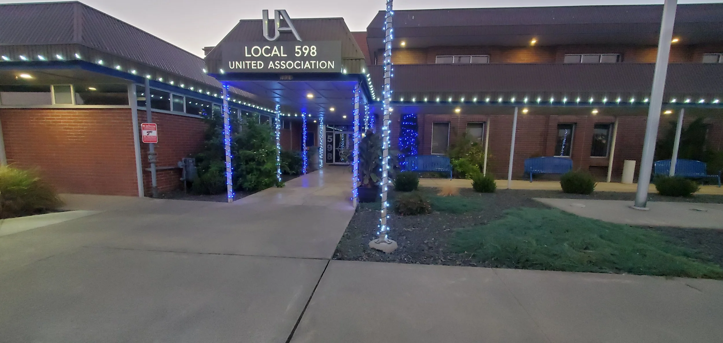 Entrance of a community building with a sign reading 'U4 Local 598 United Association,' decorated with blue string lights, with benches and shrubs outside in the evening.