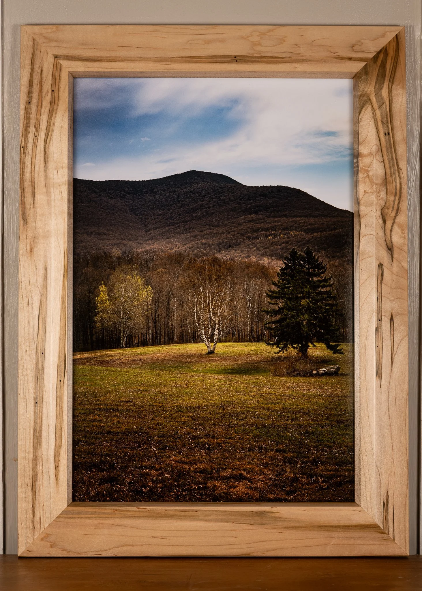 A framed landscape photograph of a mountain and forest, viewed through a wooden window frame. The scene includes a mountain with trees at the base and a partly cloudy sky.