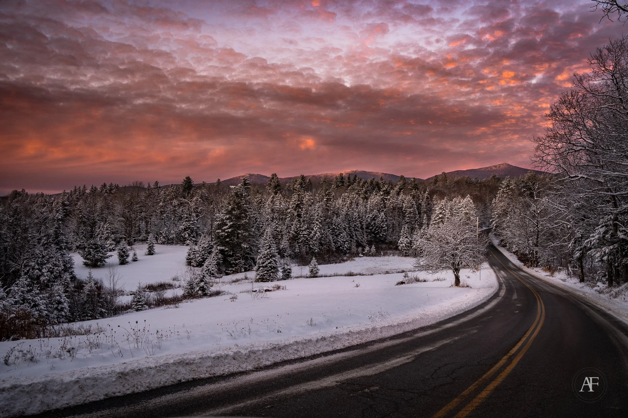 Snow-covered trees and a winding road at dusk with a colorful cloudy sky.