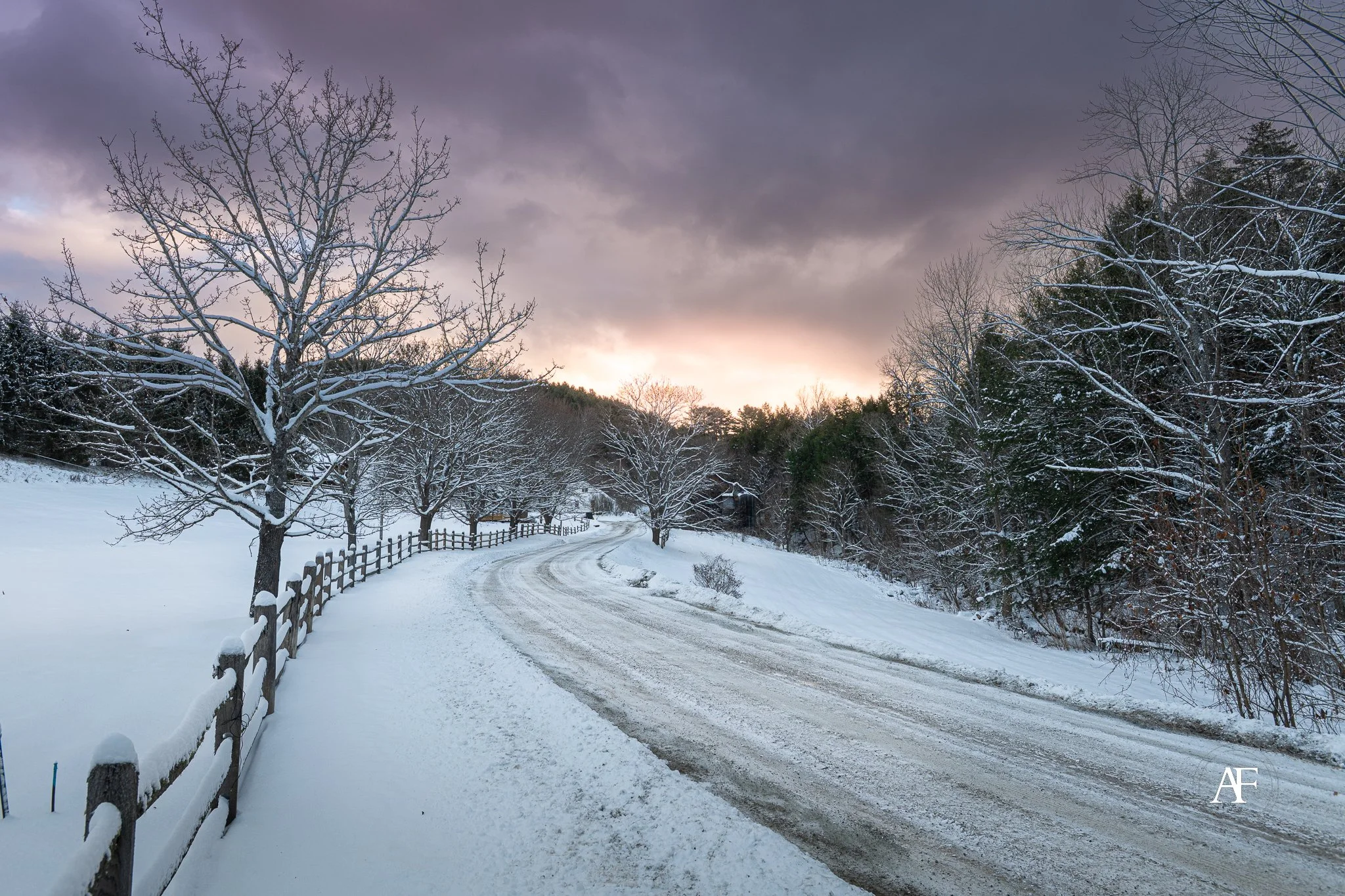 A snowy landscape with a winding dirt road, a wooden fence on the left, leafless trees on both sides, and a cloudy sky at sunset.