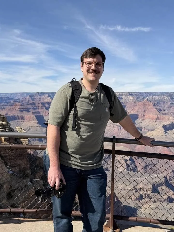 Man with glasses and mustache standing at the Grand Canyon viewpoint, holding a camera, wearing a gray shirt and backpack, during daytime with clear sky.