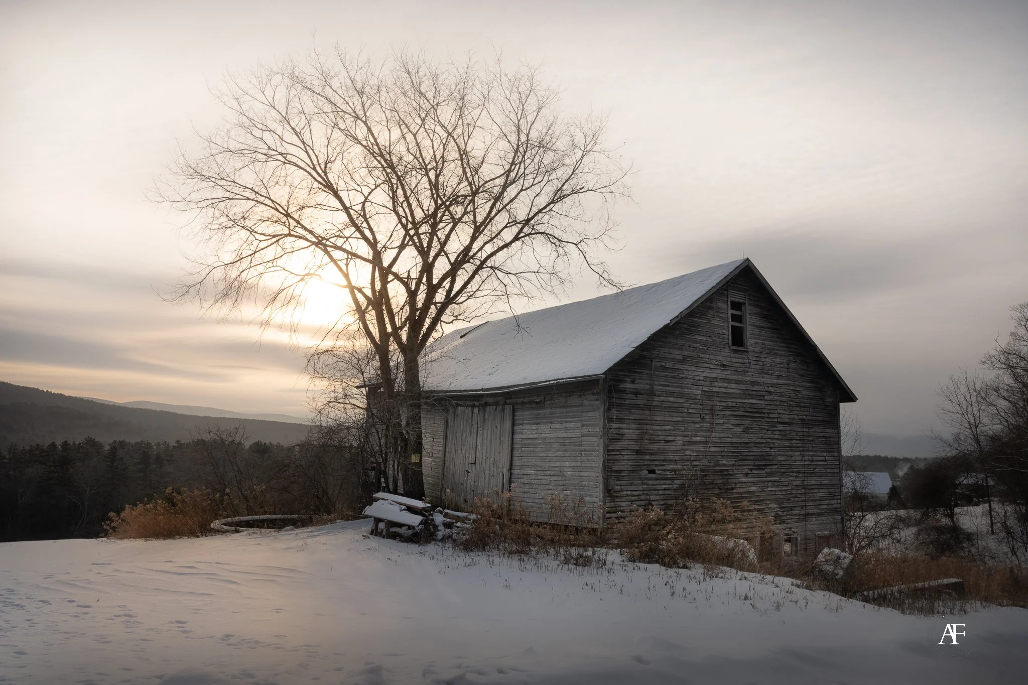 An old wooden barn next to a leafless tree during winter, with snow on the ground and a cloudy sky.