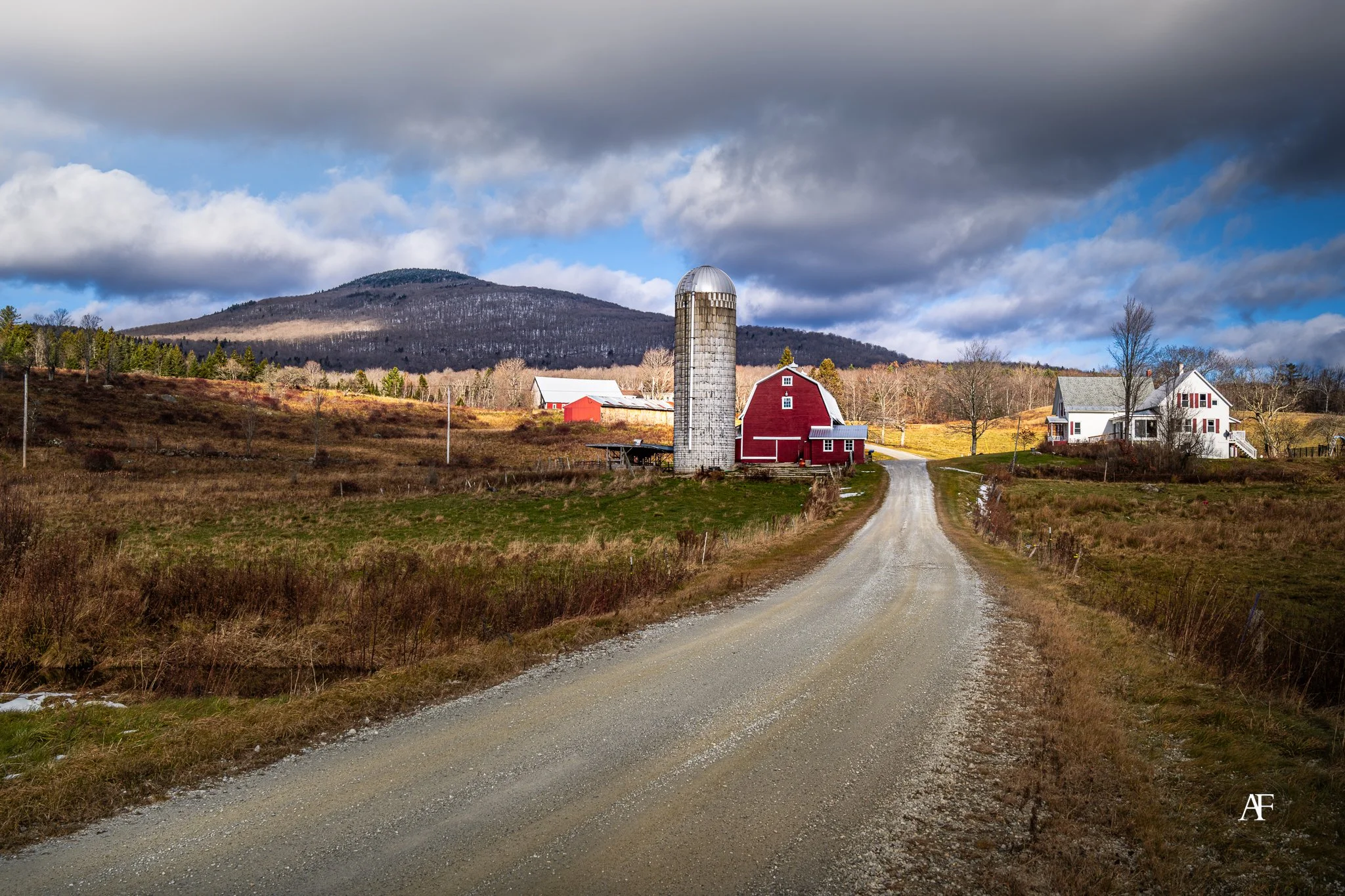 A rural landscape with a gravel road leading to farm buildings including a red barn and silo. There are trees and grassy fields, with a mountain in the background under a cloudy sky.