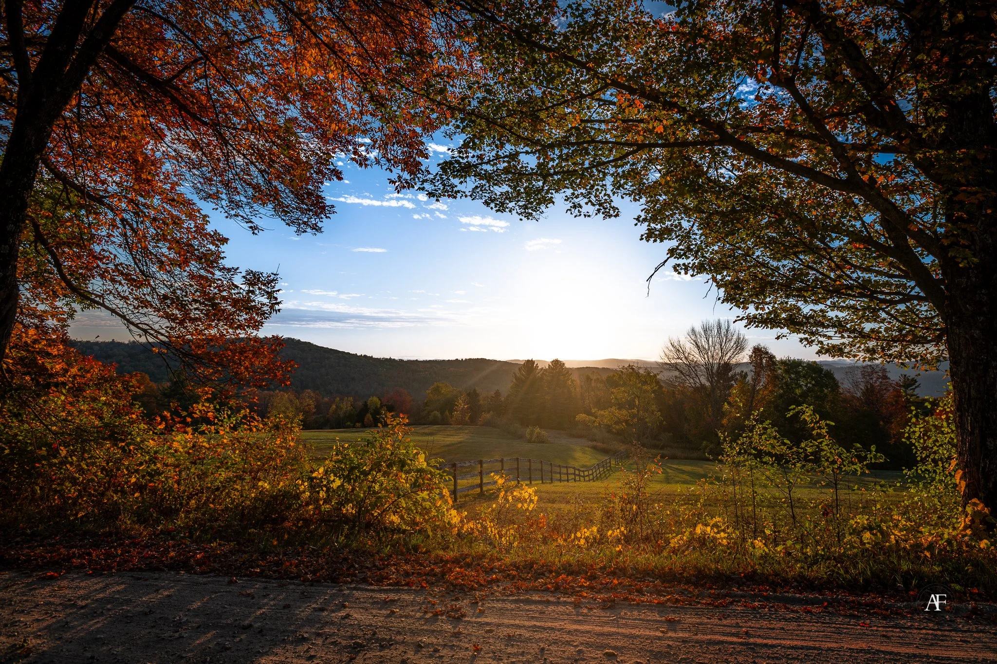 Scenic view of a countryside landscape during sunset, with trees displaying autumn foliage, a wooden fence, hills in the background, and a partly cloudy sky.