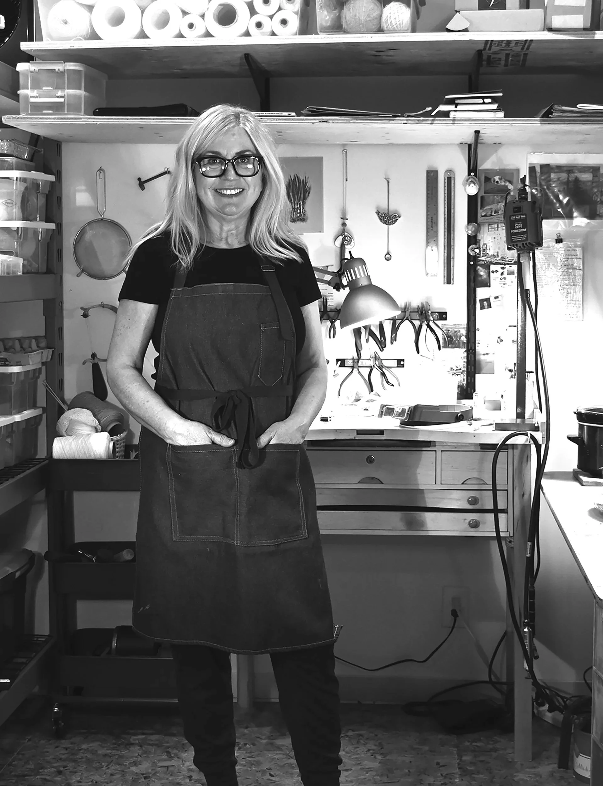 A smiling woman with blonde hair, glasses, and a black apron standing in a workshop with tools hanging on the wall and shelves filled with materials in the background.
